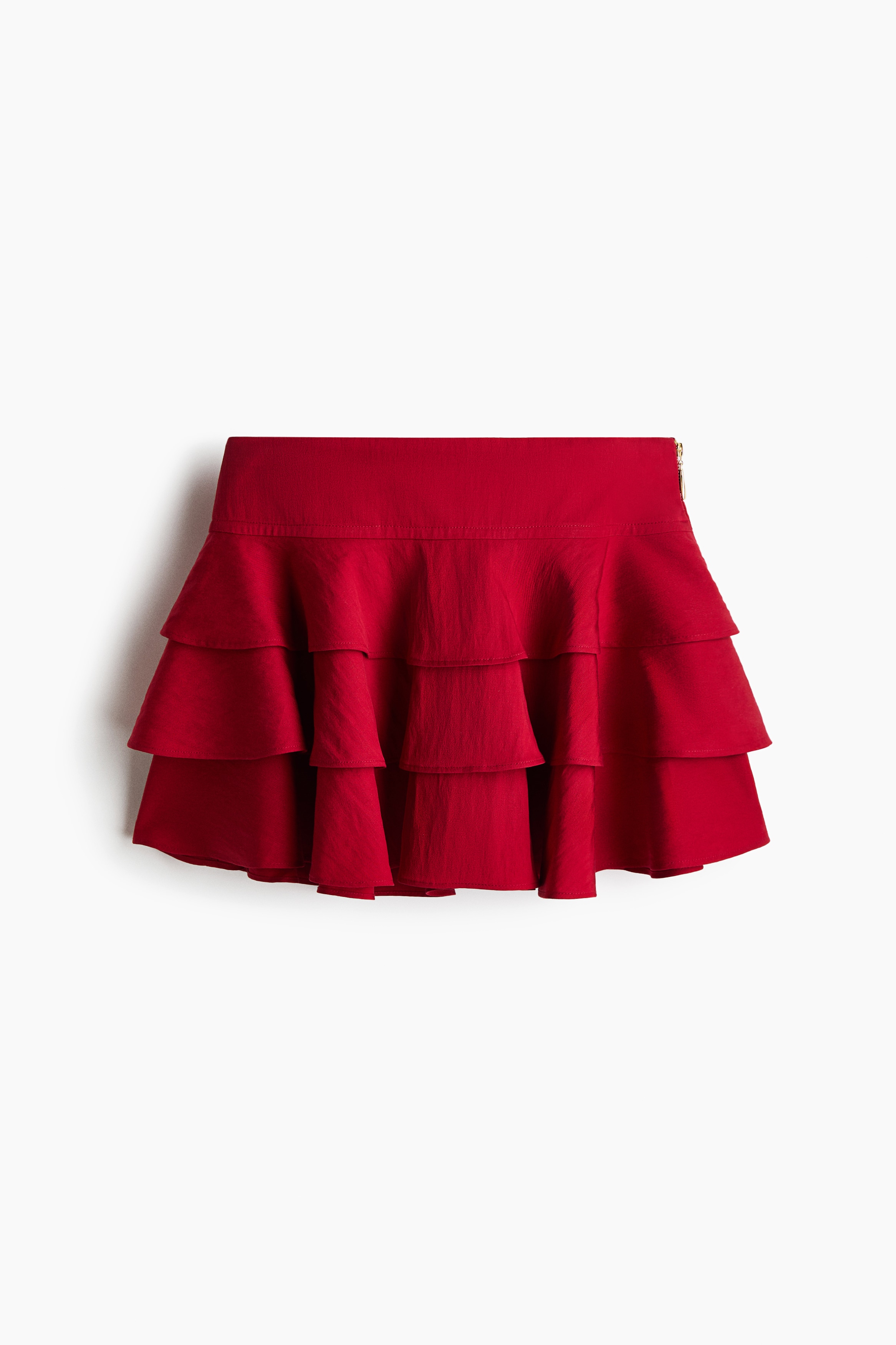 A red mini skirt featuring a double-layered waistband and three flounced tiers is being worn by the model. The skirt is paired with a matching red cropped camisole top that also features ruffled layers.