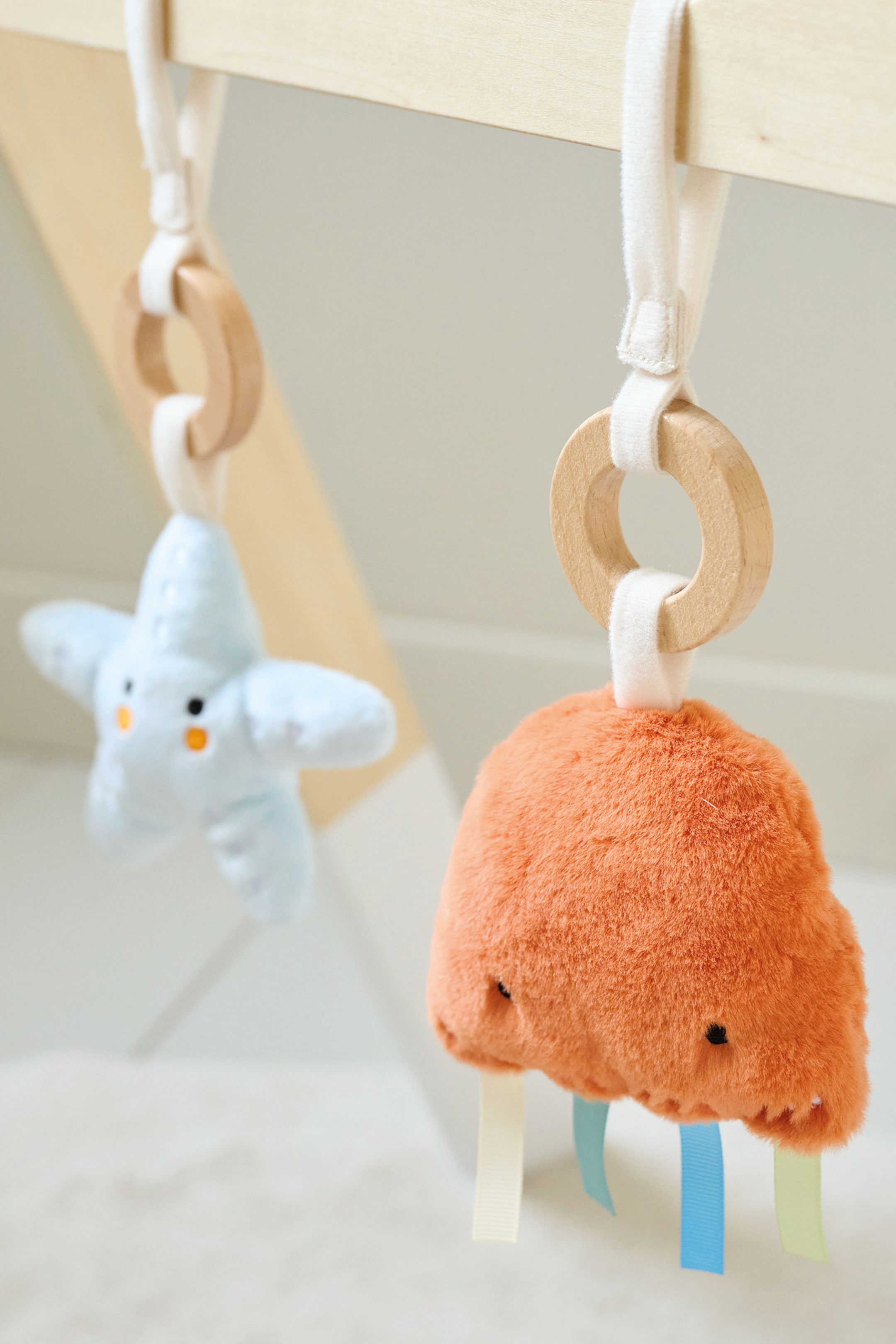 Grotere afbeelding bekijken: Twee knuffelspeeltjes voor baby's hangen aan een houten staaf. Een lichtblauw stervormig speeltje met geborduurde ogen kijkt naar voren, opgehangen aan een witte stoffen band en houten ring, terwijl rechts daarvan een oranje, onregelmatig gevormd knuffelspeeltje met geborduurde ogen en kleurrijke stoffen linten ook naar voren kijkt en op een vergelijkbare manier is bevestigd.