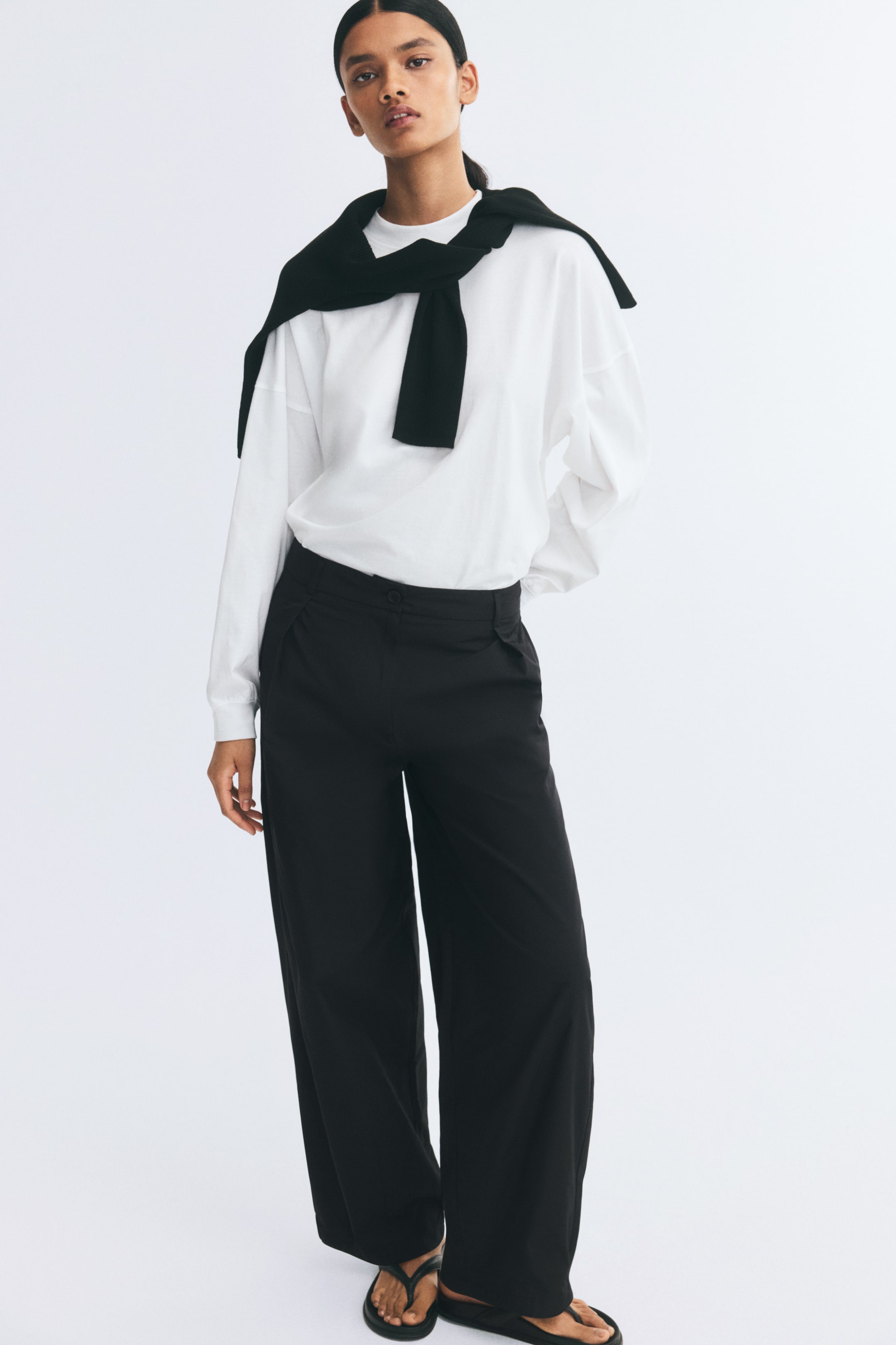 Vezi imaginea mai mare: Pantaloni negri, cu croială lejeră, din twill, cu închidere cu nasture, pense în partea de sus și crac baril, sunt purtați de model. Pantalonii sunt asortați cu un top alb cu mânecă lungă, băgat în pantaloni, și cu un pulover negru purtat pe umeri.