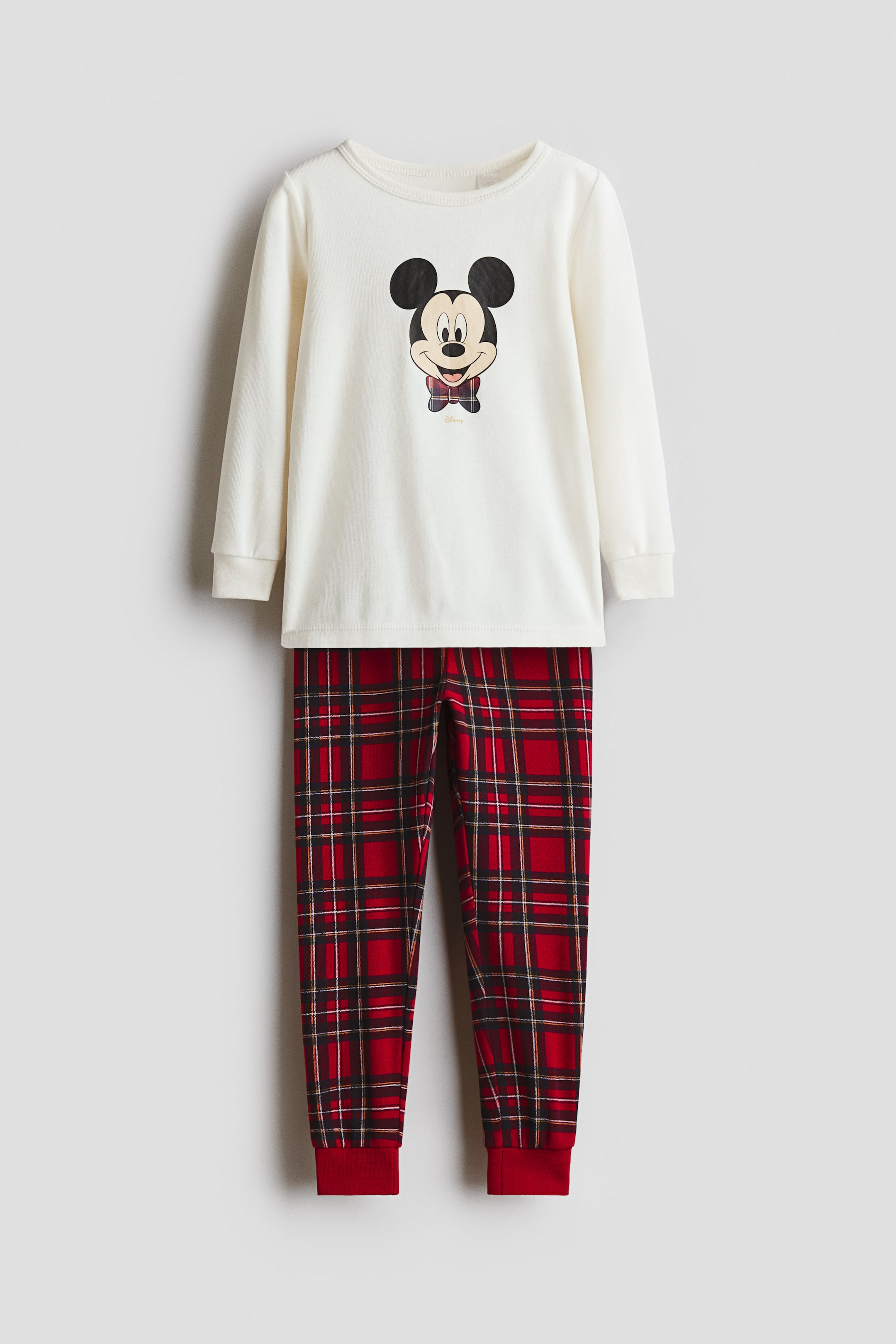 View larger image: A front-facing two-piece pajama set features a cream-colored, long-sleeved top and matching red tartan pants. The top displays a central graphic of Mickey Mouse wearing a plaid bow tie, designed with a round neckline and ribbed cuffs. The pants showcase a red, black, and white tartan pattern with solid red ribbed cuffs at the ankles.