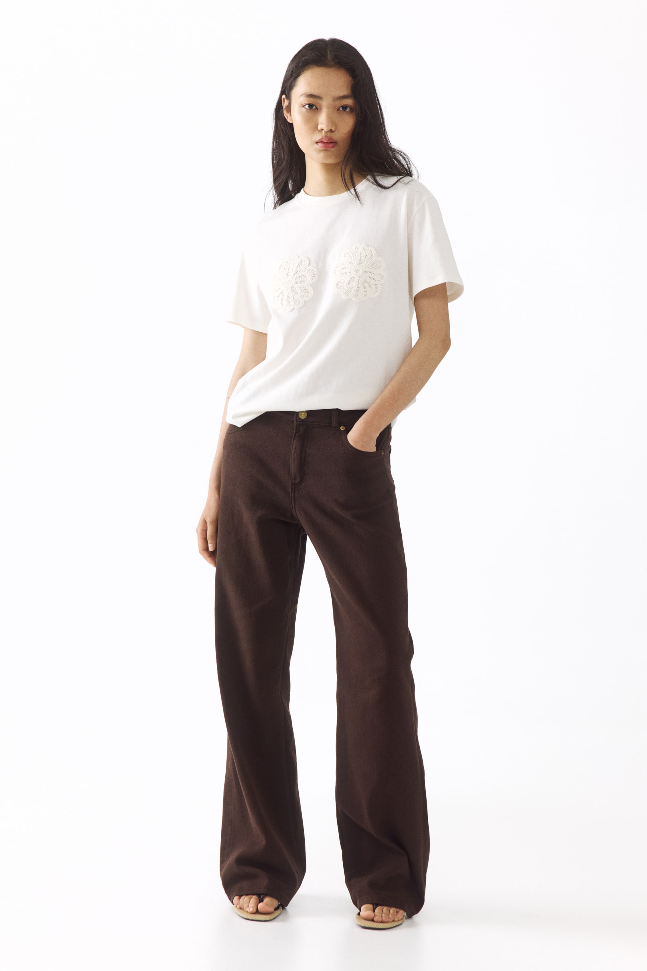 View larger image: The ensemble includes a white short-sleeved t-shirt adorned with two circular floral lace appliques on the chest, paired with brown wide-leg trousers featuring front pockets and a visible button and zipper closure.