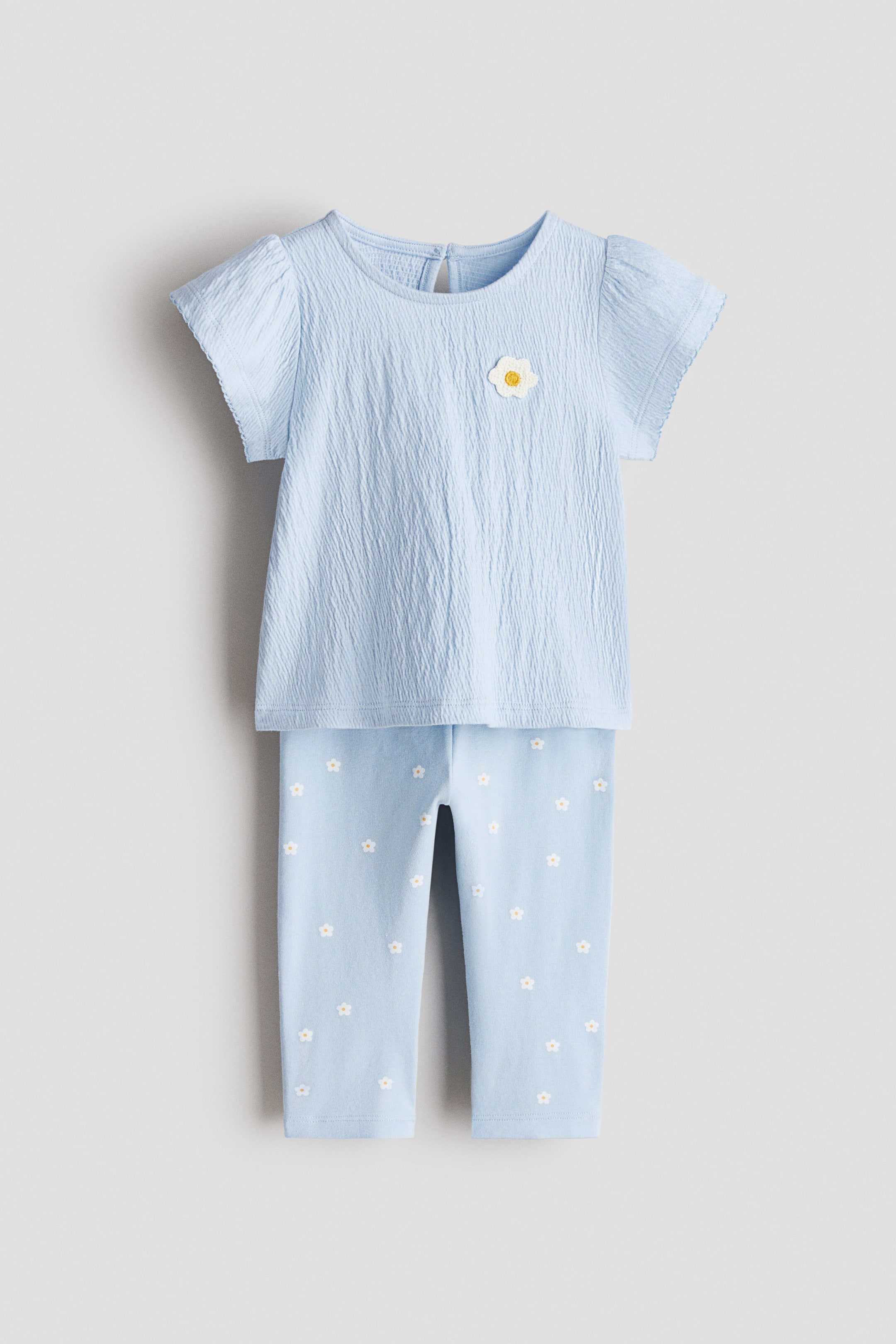 Büyük resmi görüntüle: İki parçalı takım, açık mavi bir üst ve uyumlu tayttan oluşmakta olup her ikisi önden gösterilmektedir. Açık mavi üst kırışık bir dokuya, kısa balon kollara ve yuvarlak yakaya sahiptir; sol üst göğüs kısmında beyaz ve sarı çiçek aplikesi yer alır. Eşlik eden açık mavi tayt ise küçük beyaz ve sarı çiçeklerin serpiştirildiği bir desene sahiptir.