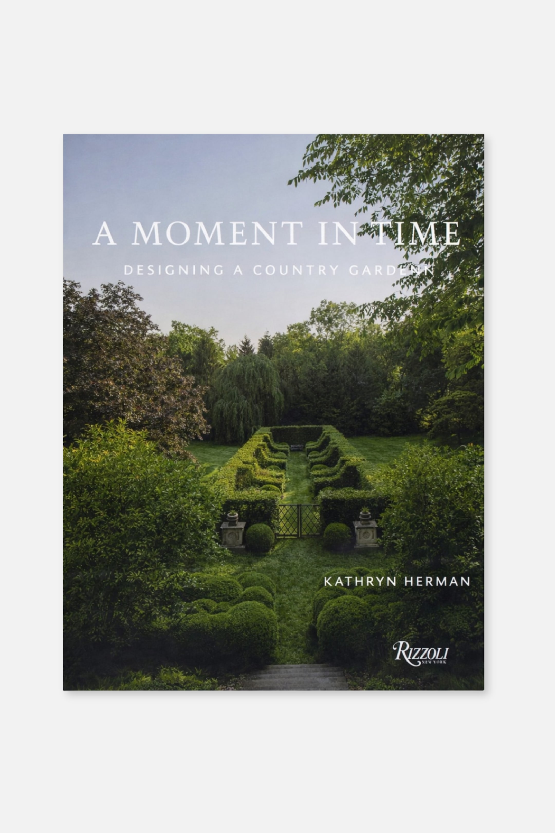 Agrandir l'image: Une couverture de livre orientée vers l'avant affiche une photographie d'un jardin à la française avec des haies vertes taillées, une grille noire et des arbres luxuriants sous un ciel bleu clair. Le titre "Un Moment dans le Temps" apparaît en haut, avec "Concevoir un Jardin de Campagne" en dessous, et "Kathryn Herman" est imprimé près du bas, accompagné du logo "Rizzoli New York".