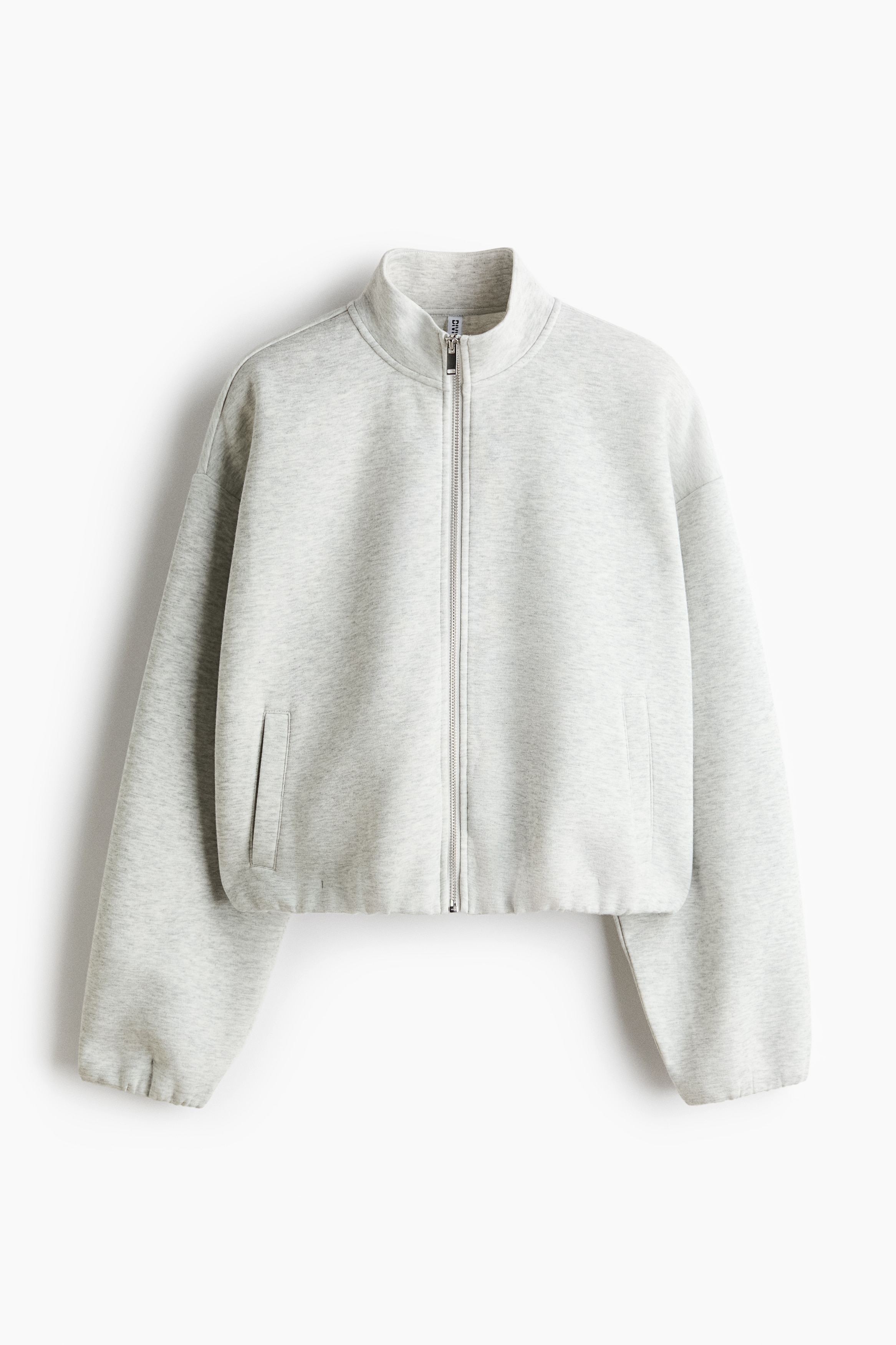 A light-grey, loose-fit scuba top is shown from the front, featuring a stand collar, a front zip, dropped shoulders and welt front pockets, with covered elastication at the cuffs and hem. The top is layered over a white ribbed crop top and paired with matching light-grey sweatpants.