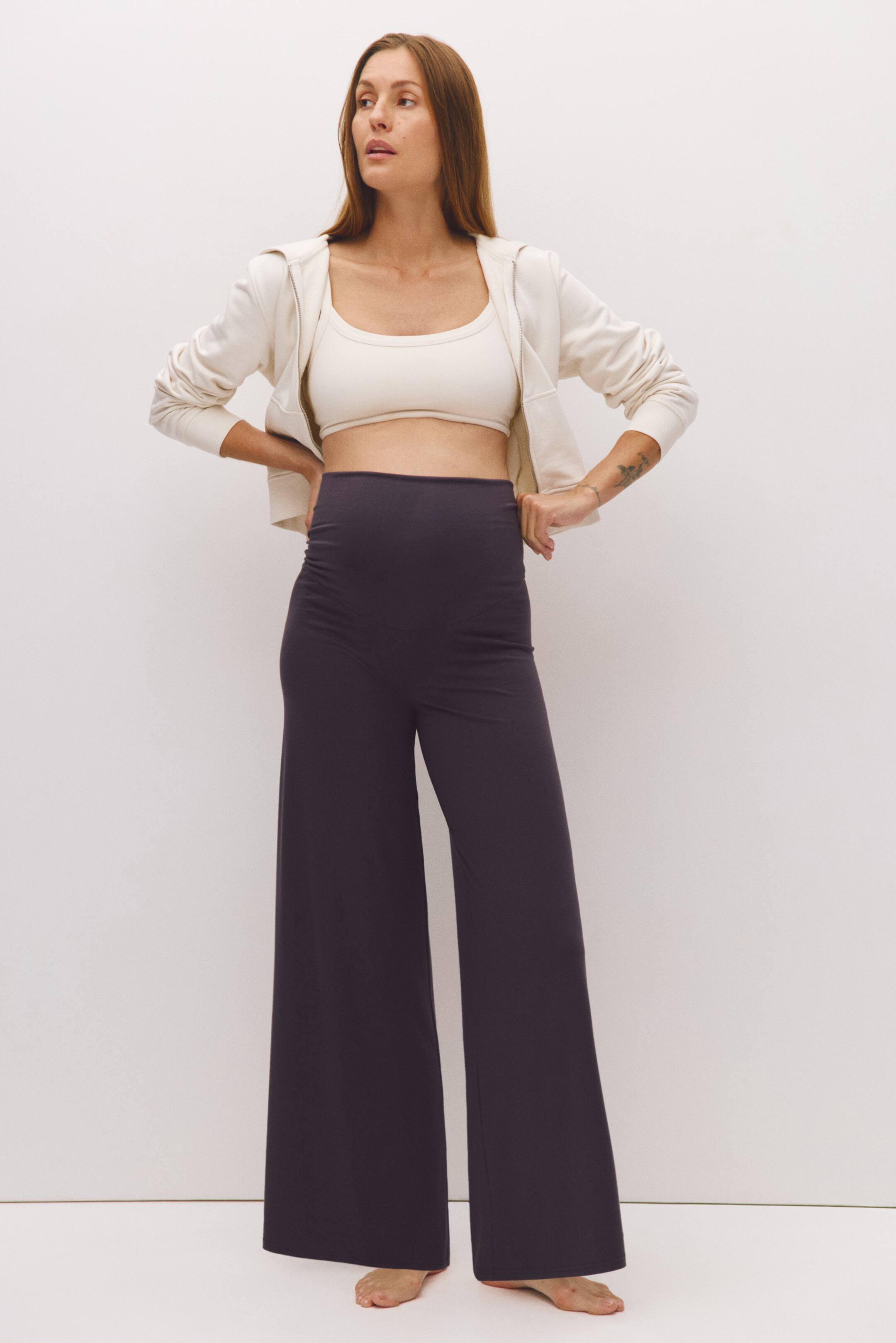 Größeres Bild ansehen: Die von dem Model getragene Yogahose aus dunkelgrauem Jersey mit hohem, weitem Bund und weitem Bein wird mit den Händen in die Taille gestützt präsentiert. Die Hose ist mit einem cremefarbenen Sport-BH und einem offenen, passenden Hoodie mit Reißverschluss kombiniert.