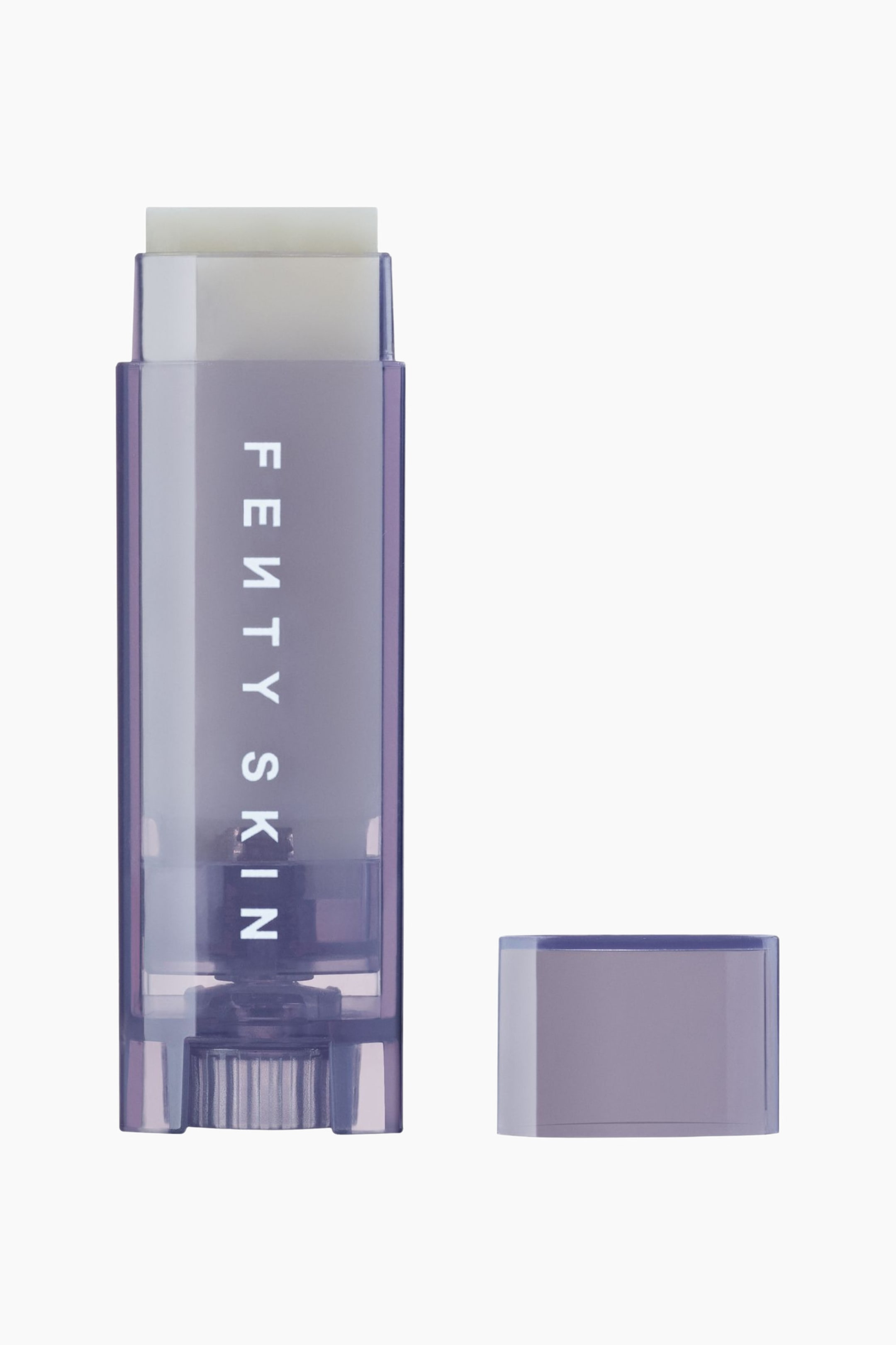 Visa större bild: Lux Balm Hydrating Moisture Barrier Lip Balm står upprätt och visar sitt halvgenomskinliga, lila tonade plastfodral med den vita logotypen "FENTY SKIN" tryckt vertikalt på framsidan. Ett fast, benvitt balm sticker ut något från tubens topp, medan den matchande lila korken ligger avtagna till höger.