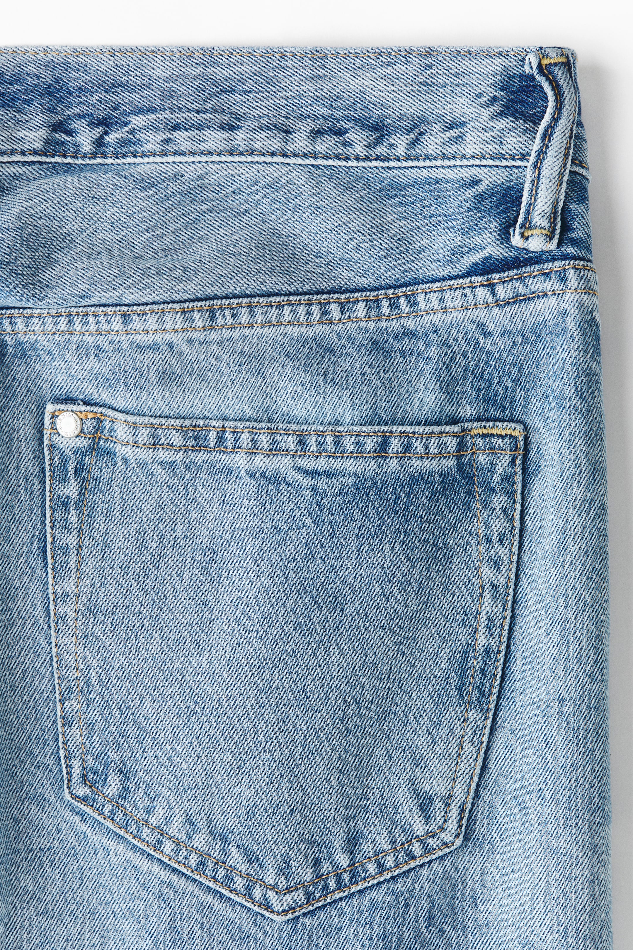 View larger image: Light blue denim jeans are showcasing their upper rear section, with a rectangular back pocket that has tan contrast stitching and a silver-toned metal rivet in the top left corner. The waistband and a single belt loop can be seen at the top.