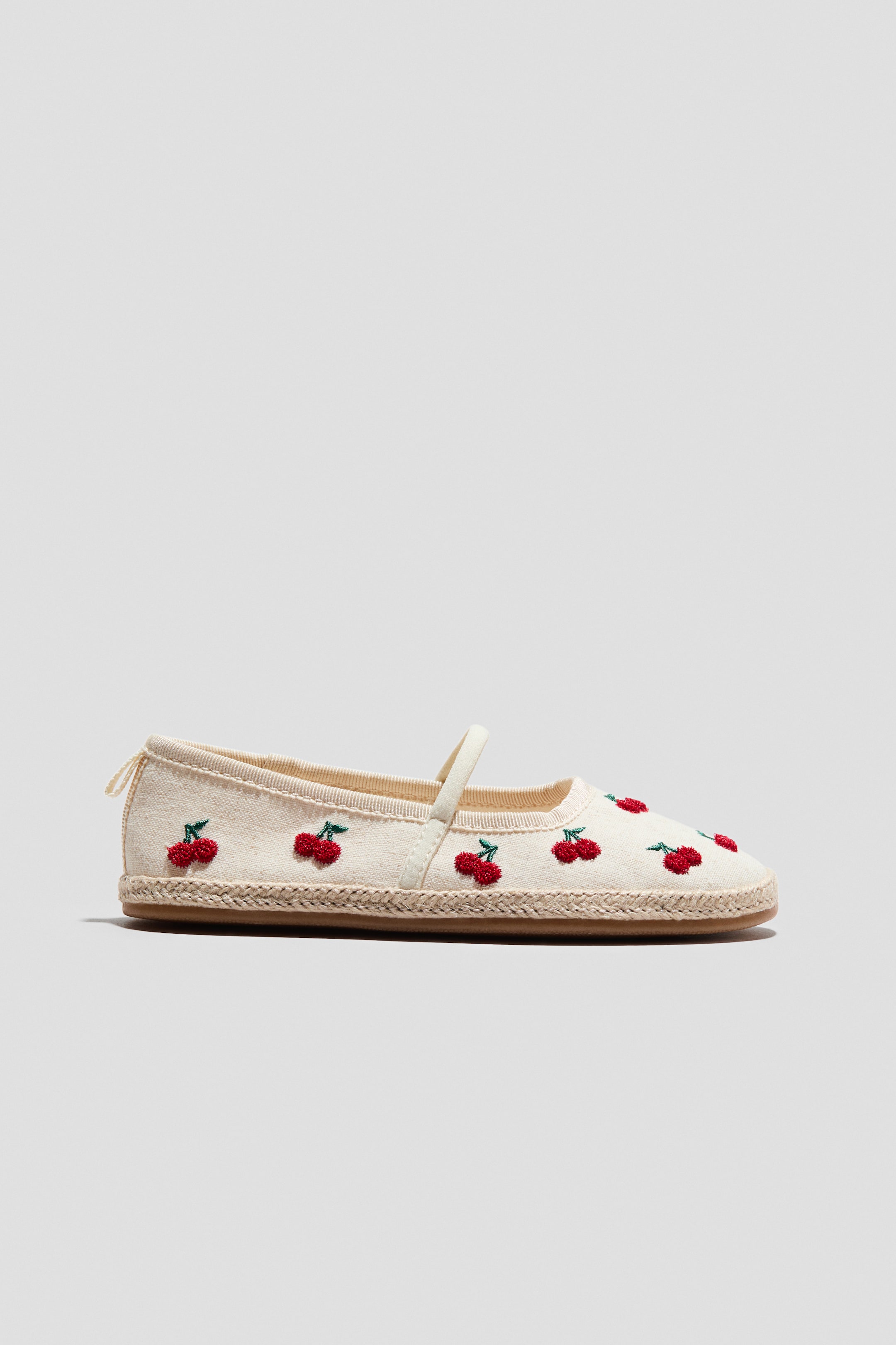Agrandir l'image: Une espadrille présente sa tige en textile de couleur crème, ornée de cerises rouges brodées. Elle se distingue par ses bouts arrondis, une fine bride élastiquée sur le pied, une petite boucle à l'arrière et une finition en jute tressé autour des semelles.