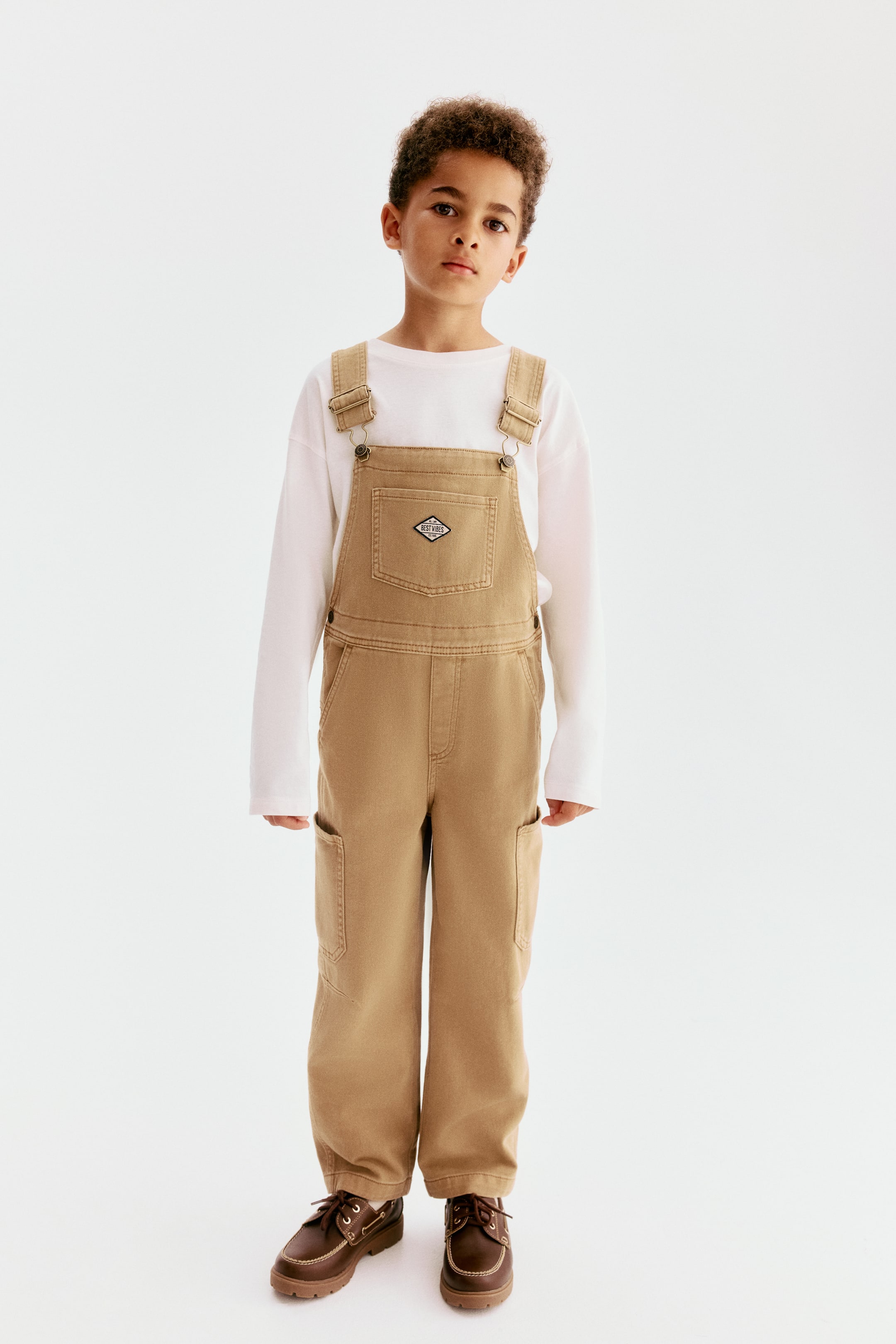 Visualizza l'immagine ingrandita: La salopette è orientata frontalmente ed è realizzata in twill di cotone lavato color tan. Presenta bretelle regolabili con fibbie metalliche, una tasca applicata sul petto con una piccola etichetta ricamata, bottoni a pressione sui lati, patta decorativa, tasche laterali diagonali e tasche applicate sulle gambe.