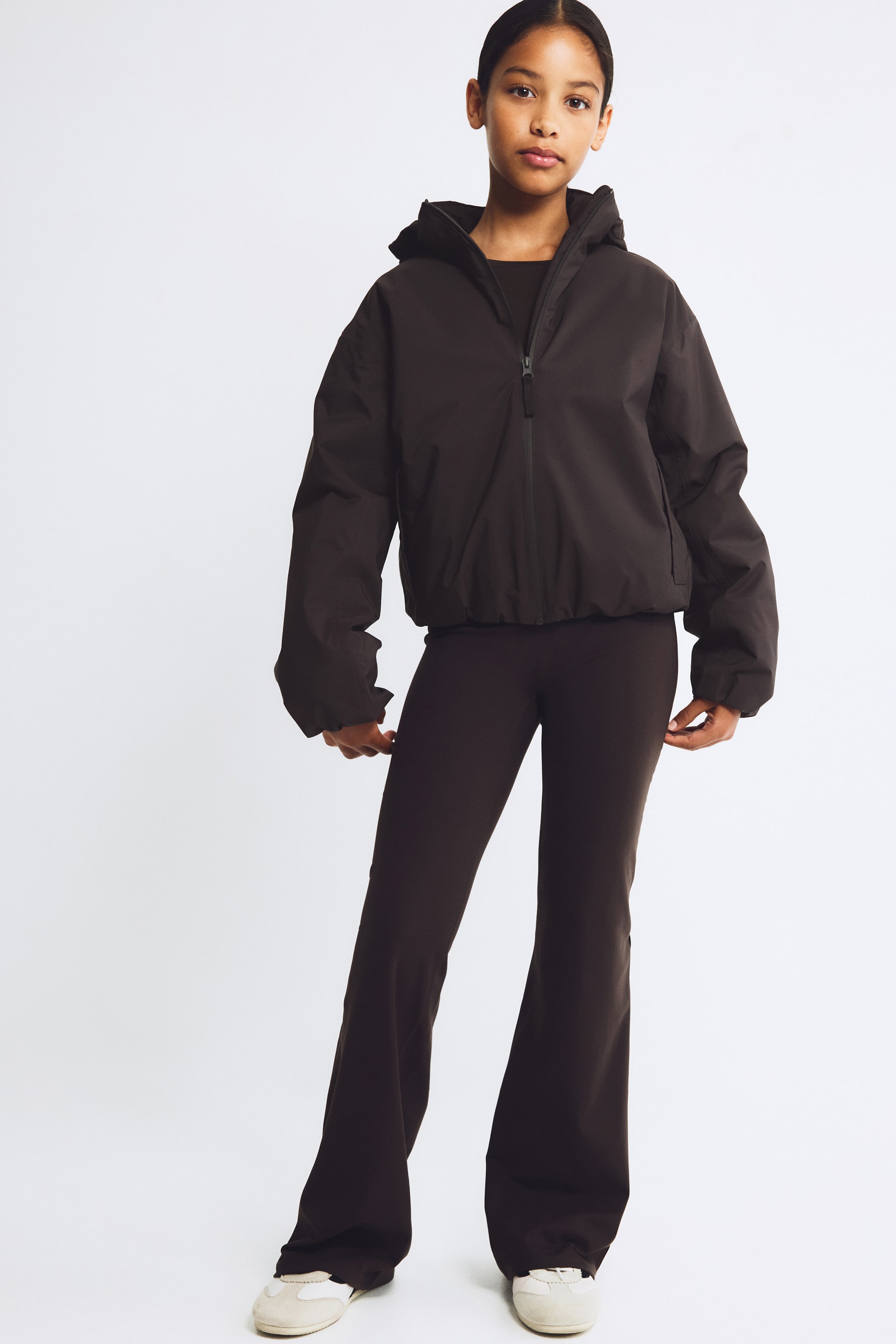 Grotere afbeelding bekijken: Een donkerbruine jas met capuchon, voorzien van een ritssluiting aan de voorkant, relaxte lange mouwen met geplooide boorden en een elastische zoom, wordt gedragen met bijpassende slim-fit uitlopende broek. Deze frontale weergave benadrukt het sportieve silhouet en de relaxte pasvorm van deze gecoördineerde set.