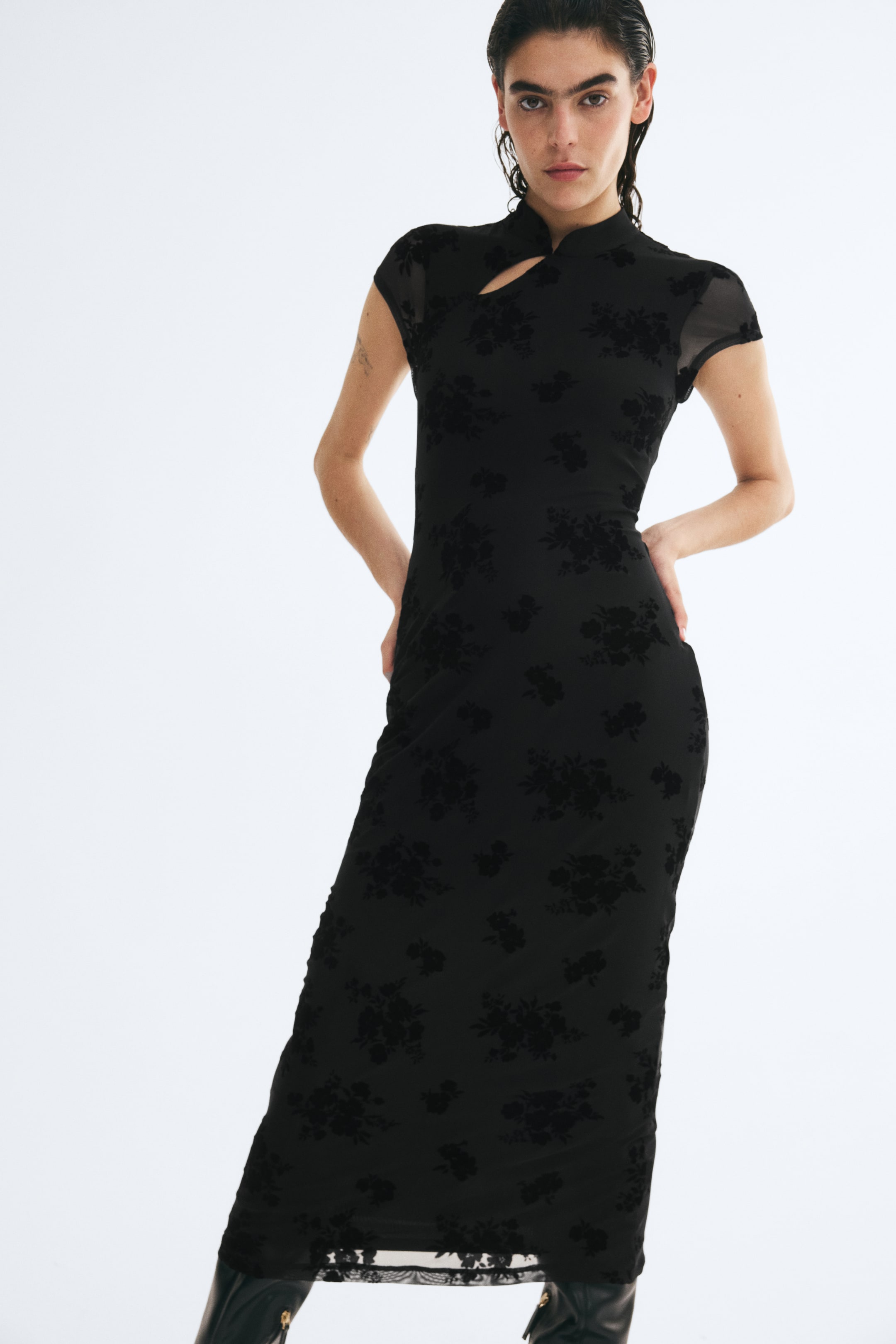 Ampliar la imagen: La modelo lleva puesto un vestido largo negro de malla ajustado con un estampado floral aterciopelado, posando con las manos en las caderas. El vestido presenta cuello alto con abertura diagonal en el escote y mangas cortas tipo casquillo.