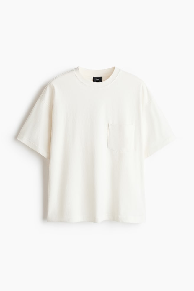 A cream-coloured t-shirt is facing forward, showcasing a crew neck and short, wide sleeves. A patch pocket is visible on the left chest. - 1