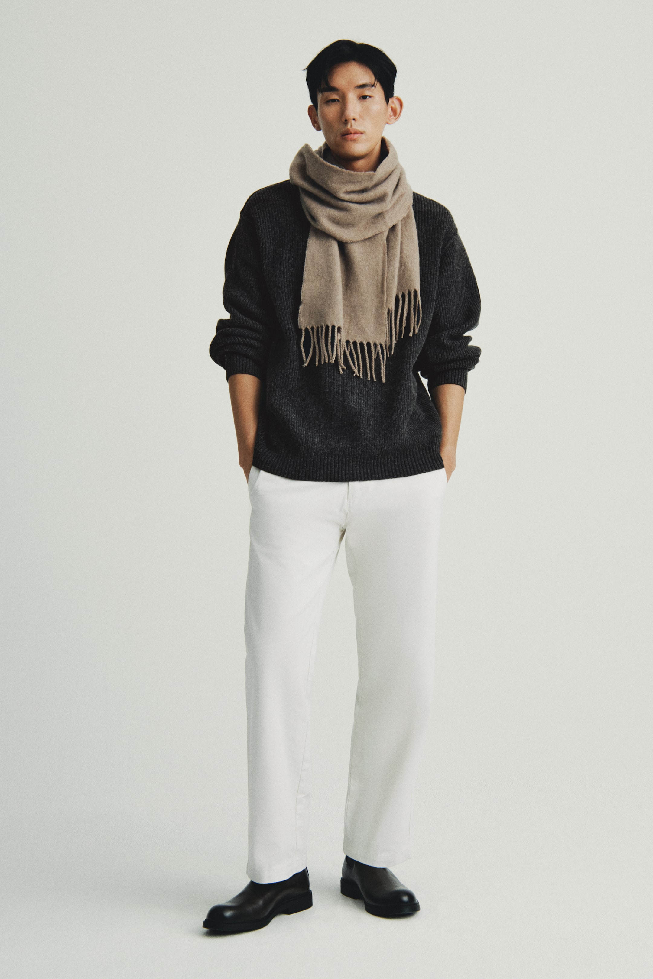 이미지 확대 보기: 짙은 회색 굵은 니트 스웨터는 흰색의 스트레이트 레그 바지와 짙은 갈색 슬립온 신발과 함께 앞쪽에서 보이도록 입고 있습니다. 밝은 갈색의 프린지 스카프는 스웨터 위에 레이어링 되며 목에 감고 있습니다.