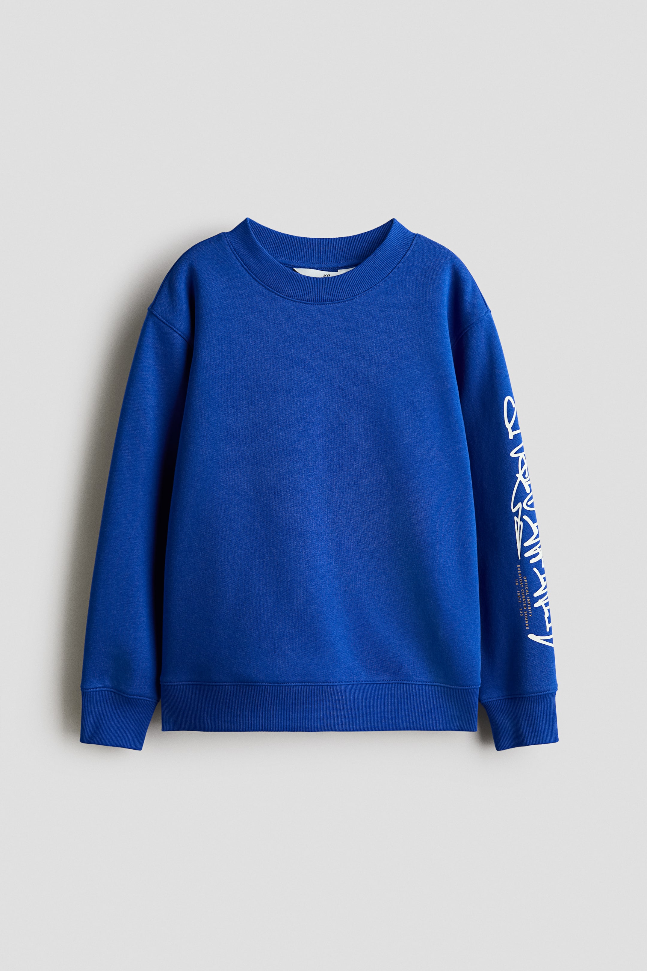 Grotere afbeelding bekijken: Een blauwe sweatshirt toont de voorkant, met een ronde, met ribboord afgewerkte halslijn, ingezakte lange mouwen en witte printmotieven op de rechtermouw. Ribboord is zichtbaar aan de manchetten en de zoom.