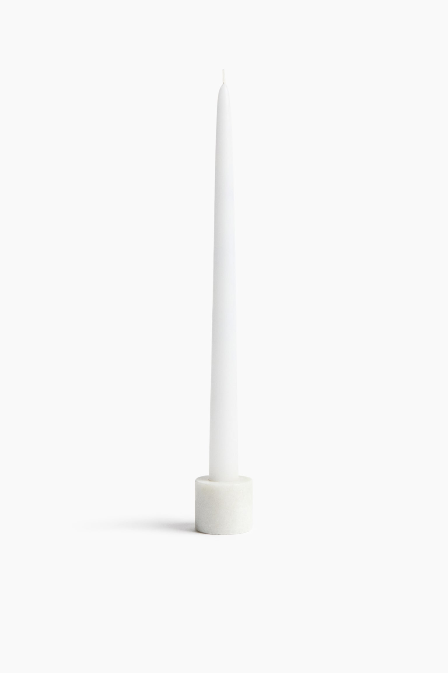 A tall, white tapered candle is standing upright, tapering to a fine point with a visible wick at the top. The candle is placed inside a small, cylindrical white marble holder with subtle grey veining.