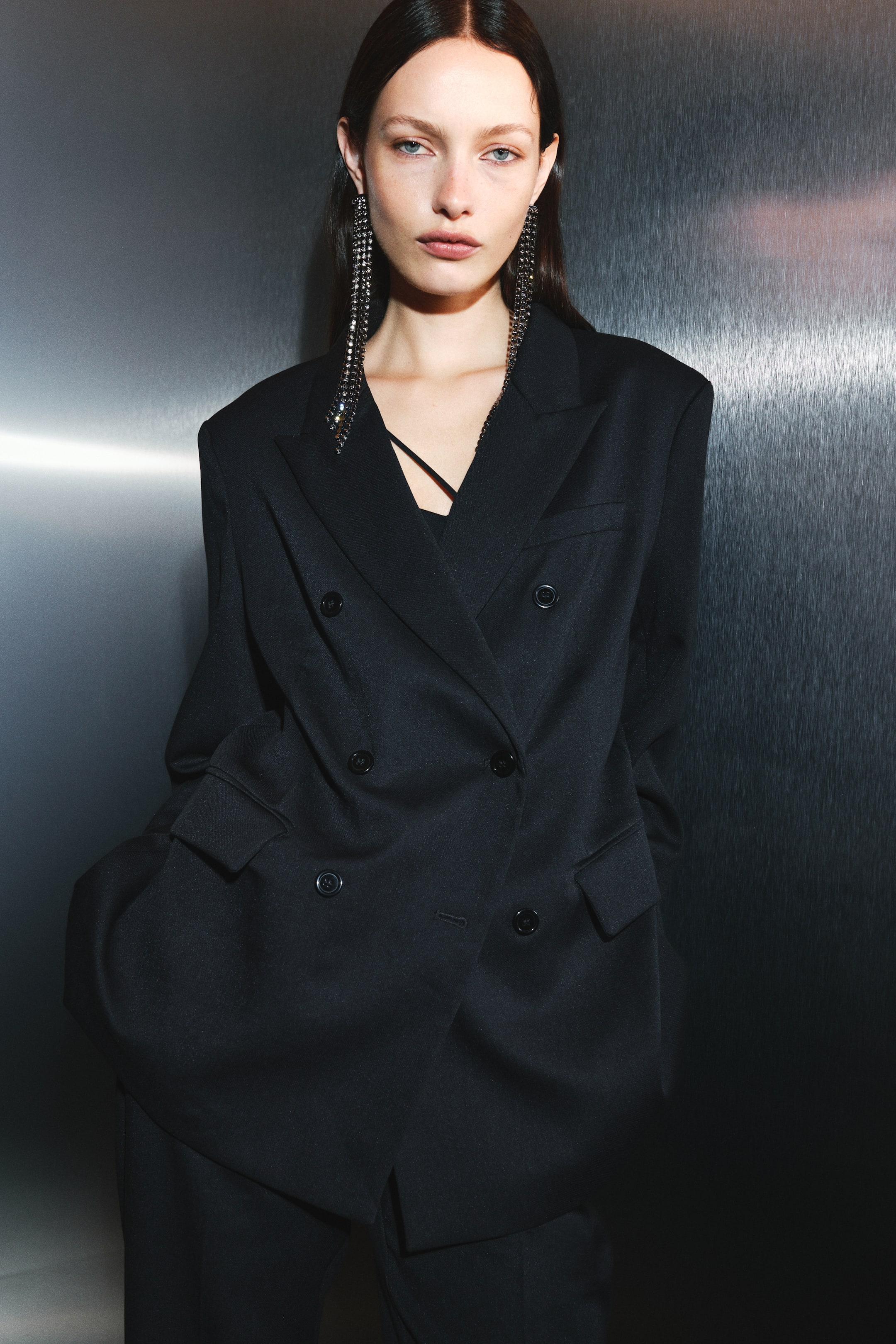 View larger image: A long, black, double-breasted blazer featuring peak lapels, structured shoulders, a welt chest pocket, and flapped front pockets is being worn by the model. The blazer is buttoned at the front and styled with matching black trousers.