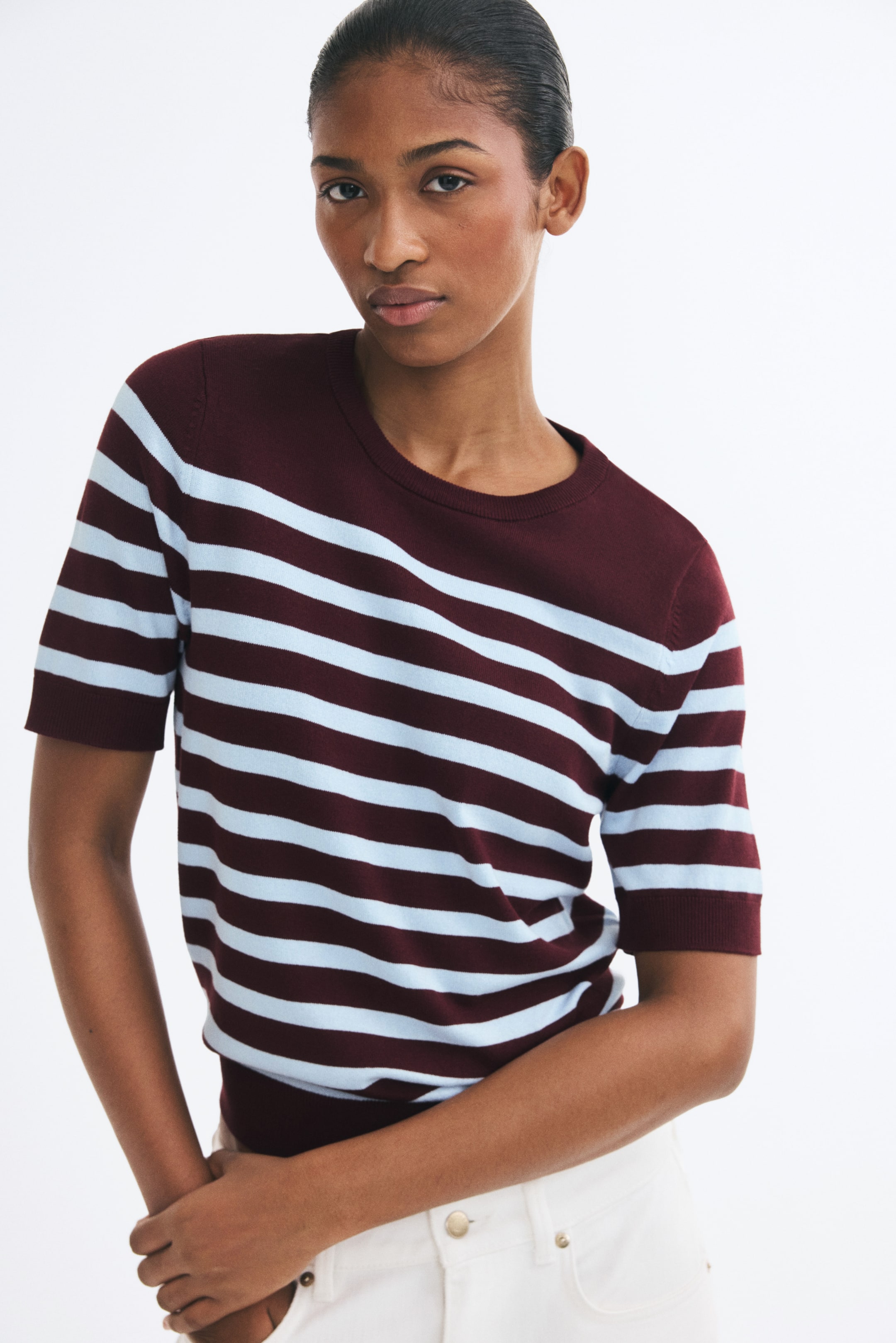 View larger image: A fine-knit, short-sleeved sweater featuring burgundy and light blue horizontal stripes, a ribbed crew neckline, and ribbed cuffs is worn by the model. The sweater is tucked into white trousers, with the model’s hands resting near the pockets.