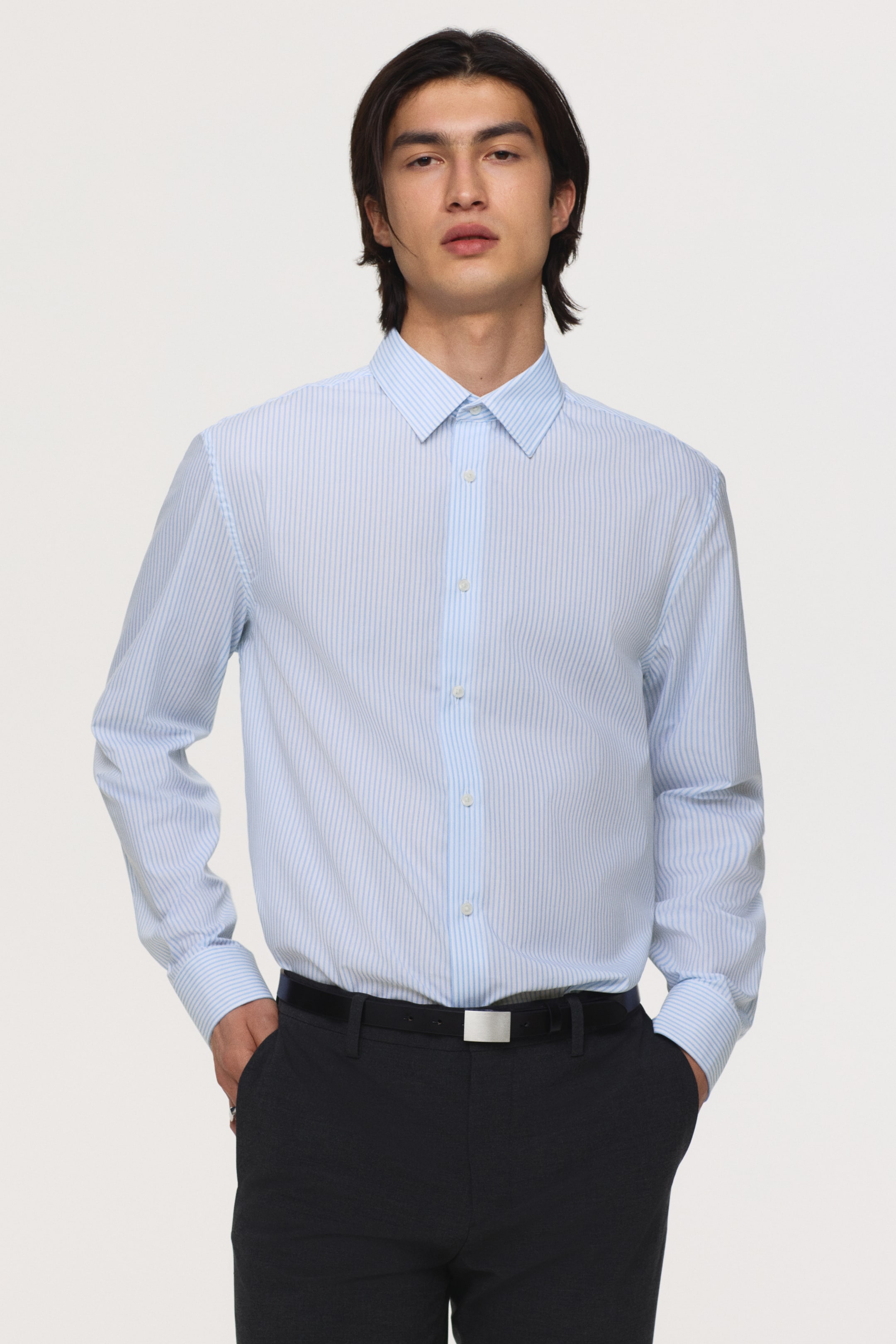 拡大画像を表示: ライトブルーの縦縞のボタンダウンシャツが前から表示されており、尖った襟とボタン付きカフスの長袖が特徴です。濃い色のパンツにインされています。
