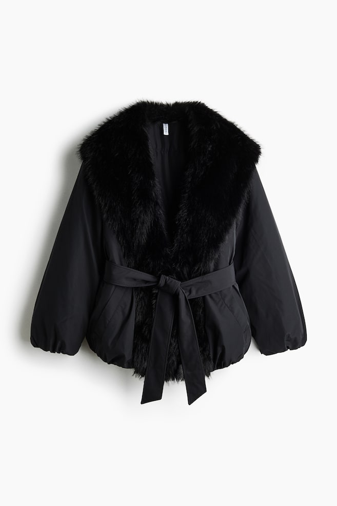 A black padded jacket is shown front-on, featuring a voluminous black faux-fur collar that extends down the front opening. The jacket is cinched at the waist with a matching self-tie fabric belt and showcases long sleeves with gathered cuffs and a puffed hem. - 1