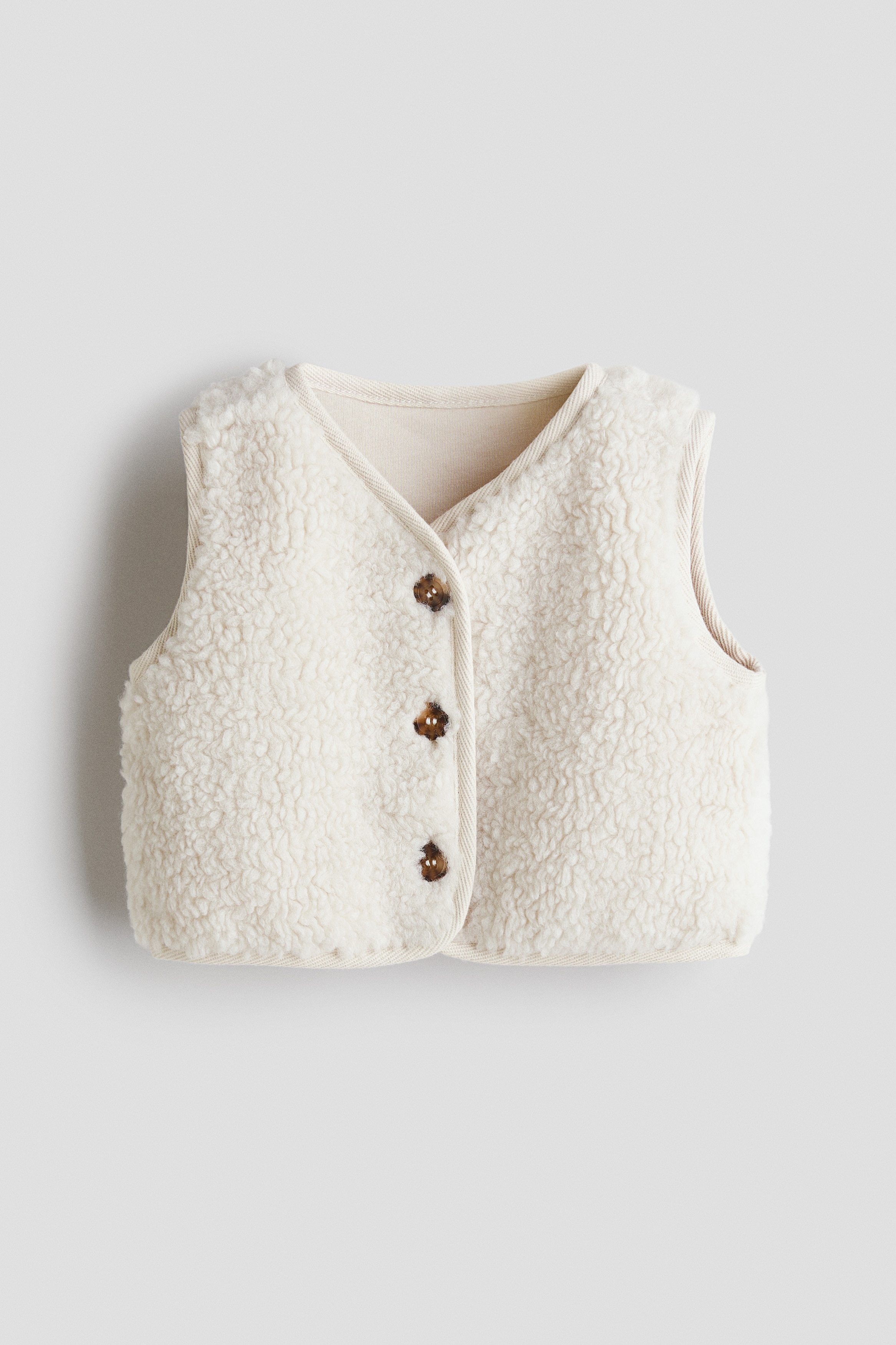 Un gilet en teddy crème doux est présenté de face, avec un col arrondi et des boutons marron foncé sur le devant. Un passepoil en twill souligne ses bords et sa patte de boutonnage, avec un haut blanc à manches longues porté en dessous.