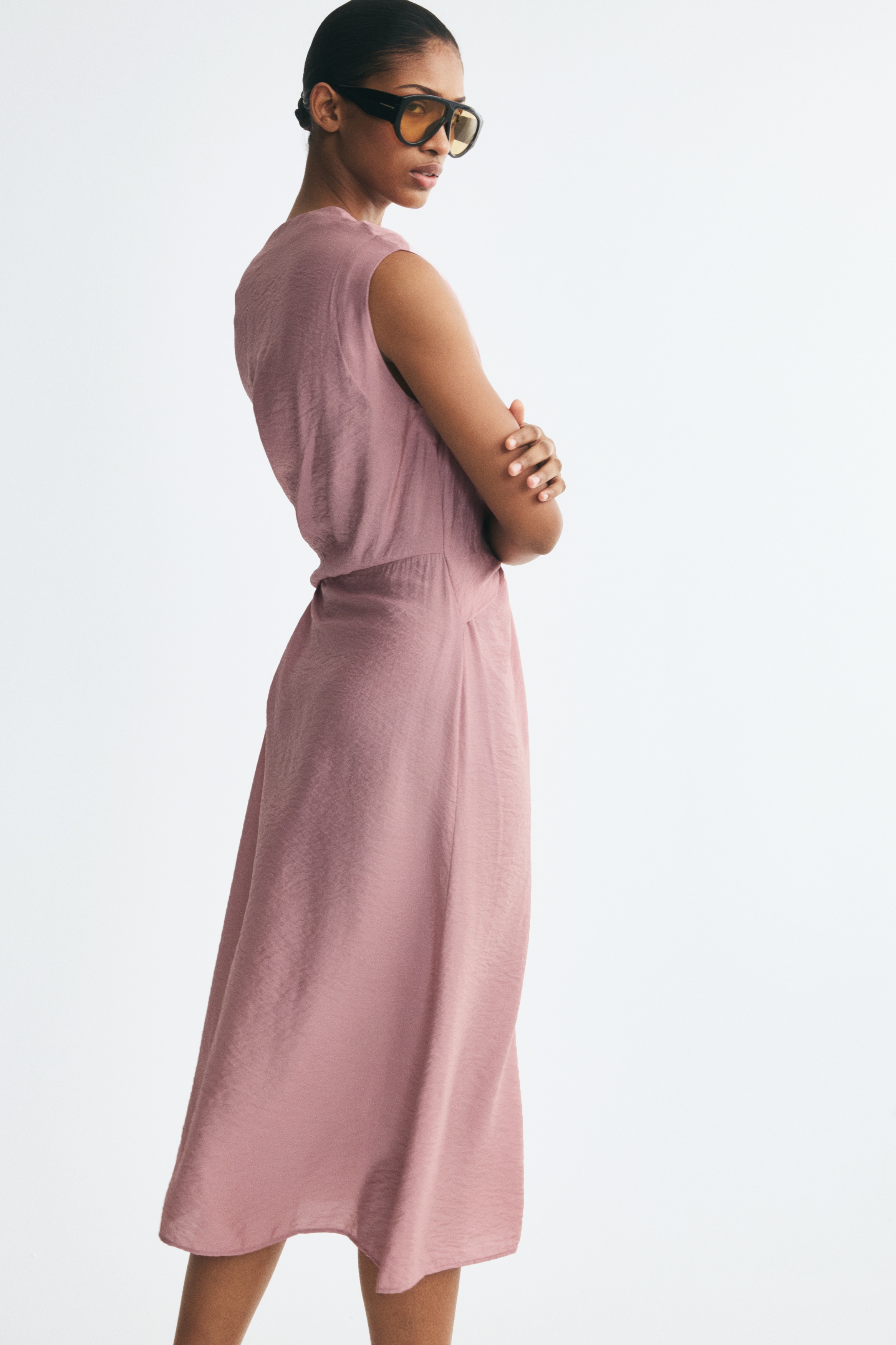 A front-facing view of a dusty rose midi dress featuring a V-neckline and cap sleeves. The dress showcases a wrap-style front overlay that ties into a knot at the waist, with the fabric draping into a fluid skirt with a subtly crinkled texture.