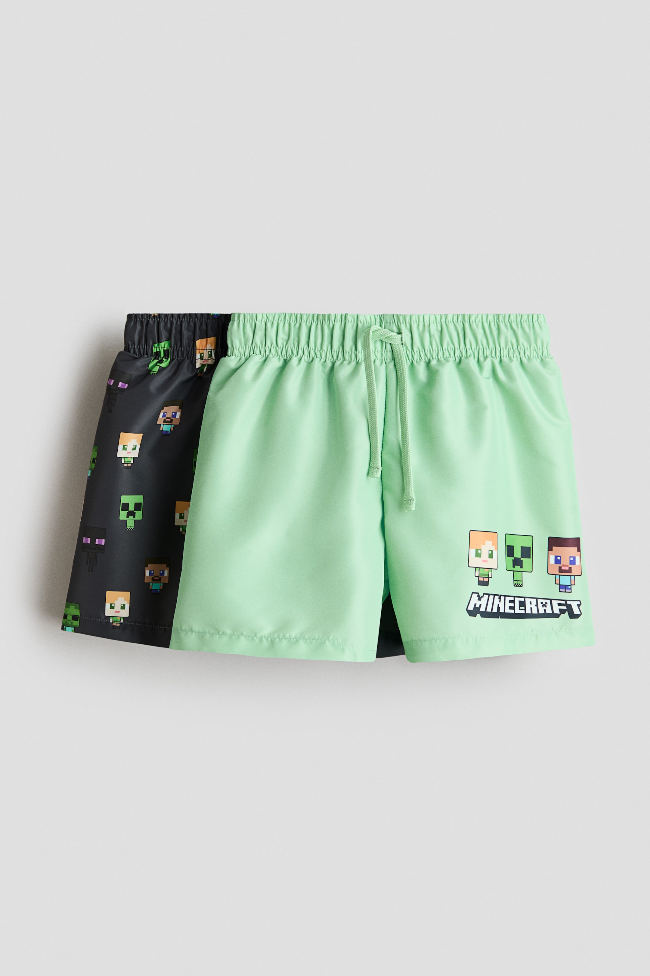 Ver imagen más grande: Se muestran dos pares de bañadores estampados, uno en verde claro y otro en gris oscuro. El par verde claro, orientado hacia el frente, presenta una cintura elástica con cordón ajustable y un estampado de personajes de Minecraft con el logo en la parte inferior de la pierna derecha. El par gris oscuro, parcialmente visible y colocado detrás, exhibe un patrón de caras de personajes de Minecraft por toda la prenda.