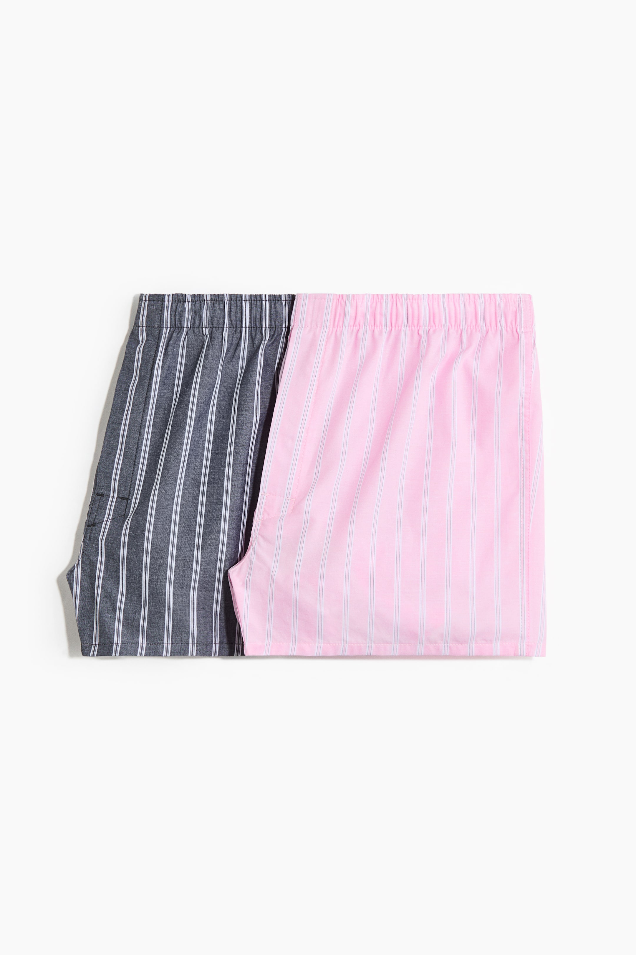 View larger image: Two pairs of boxer shorts in a soft weave are neatly laid out, showcasing their front views. The grey pair is adorned with white vertical stripes and features a button fly, while the pink pair also displays white vertical stripes. Both pairs come with elasticated waists and mid-length legs, ensuring comfort and style.