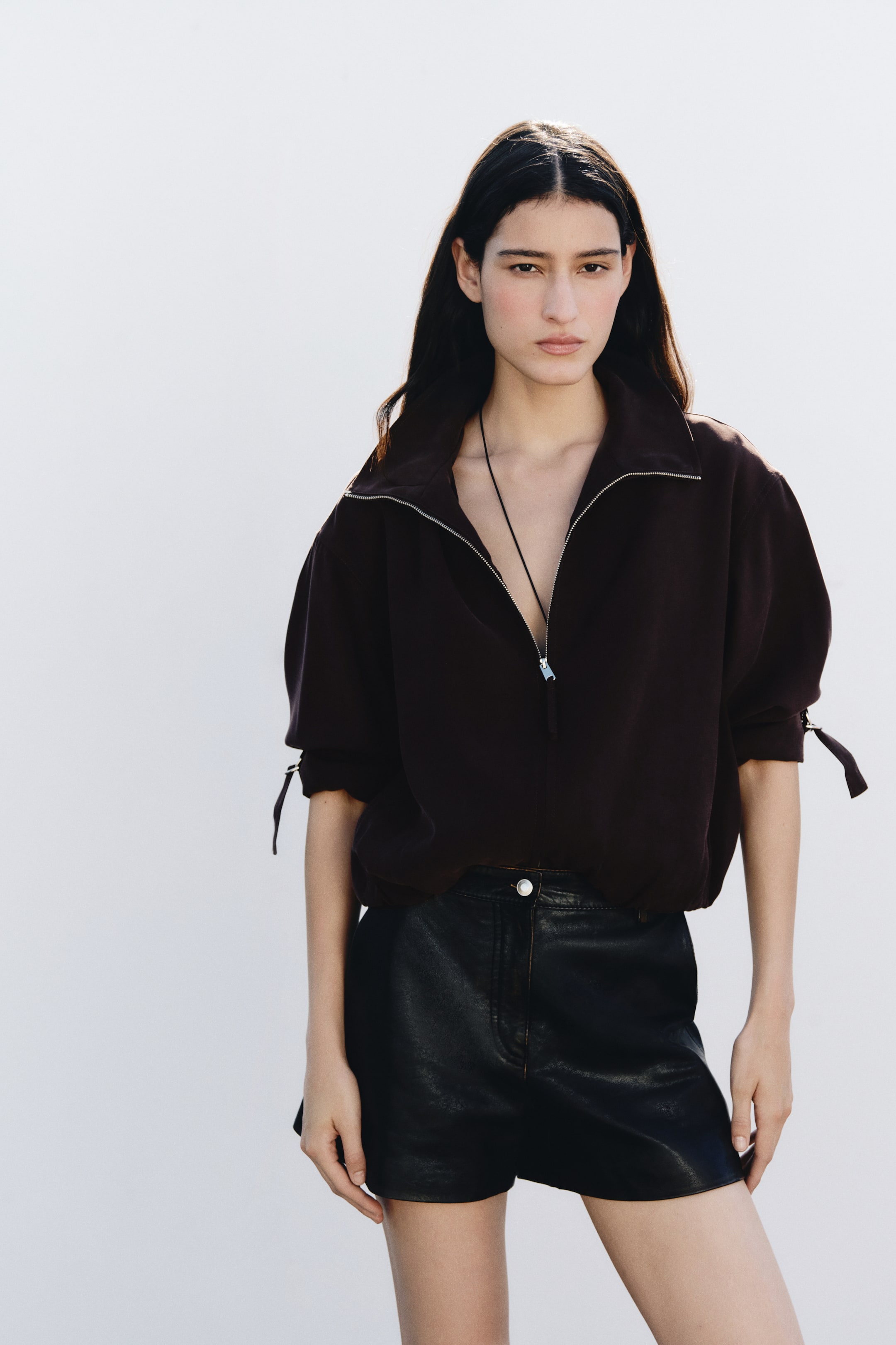 View larger image: The model is wearing a short, oversized dark brown jacket featuring a partially unzipped funnel collar, dropped shoulders, and gathered sleeves secured with tabs. The jacket is paired with black leather shorts.