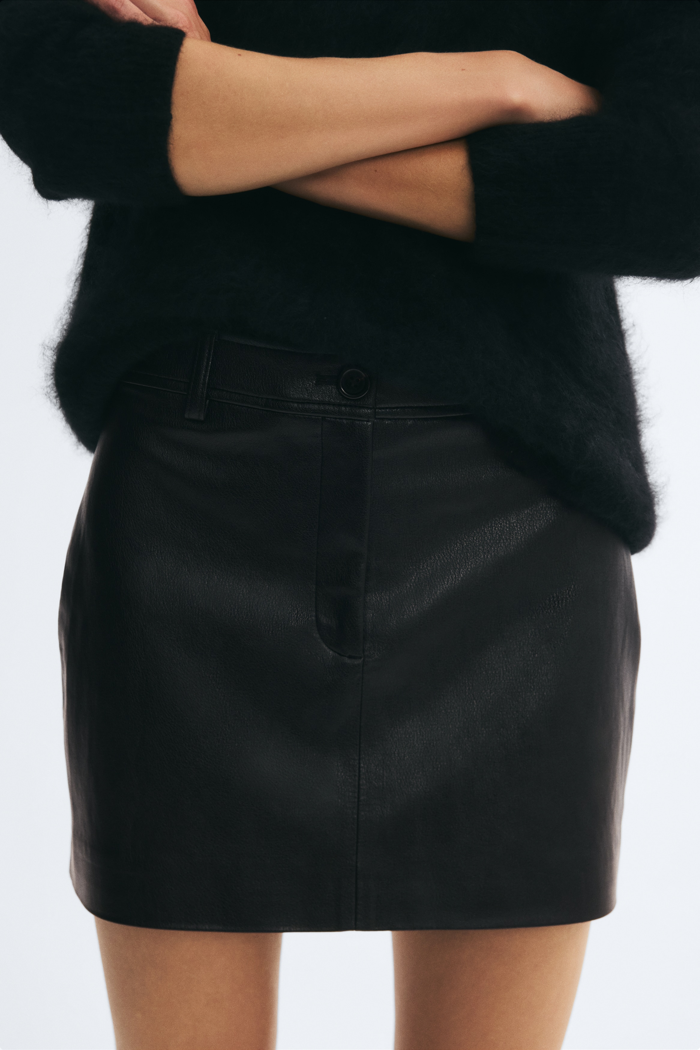 A black leather mini skirt is facing front, showcasing a structured A-line silhouette with a subtly textured finish. It features a fitted waistband with belt loops, a central button fastening, a concealed zip fly, and a vertical seam running down the center.