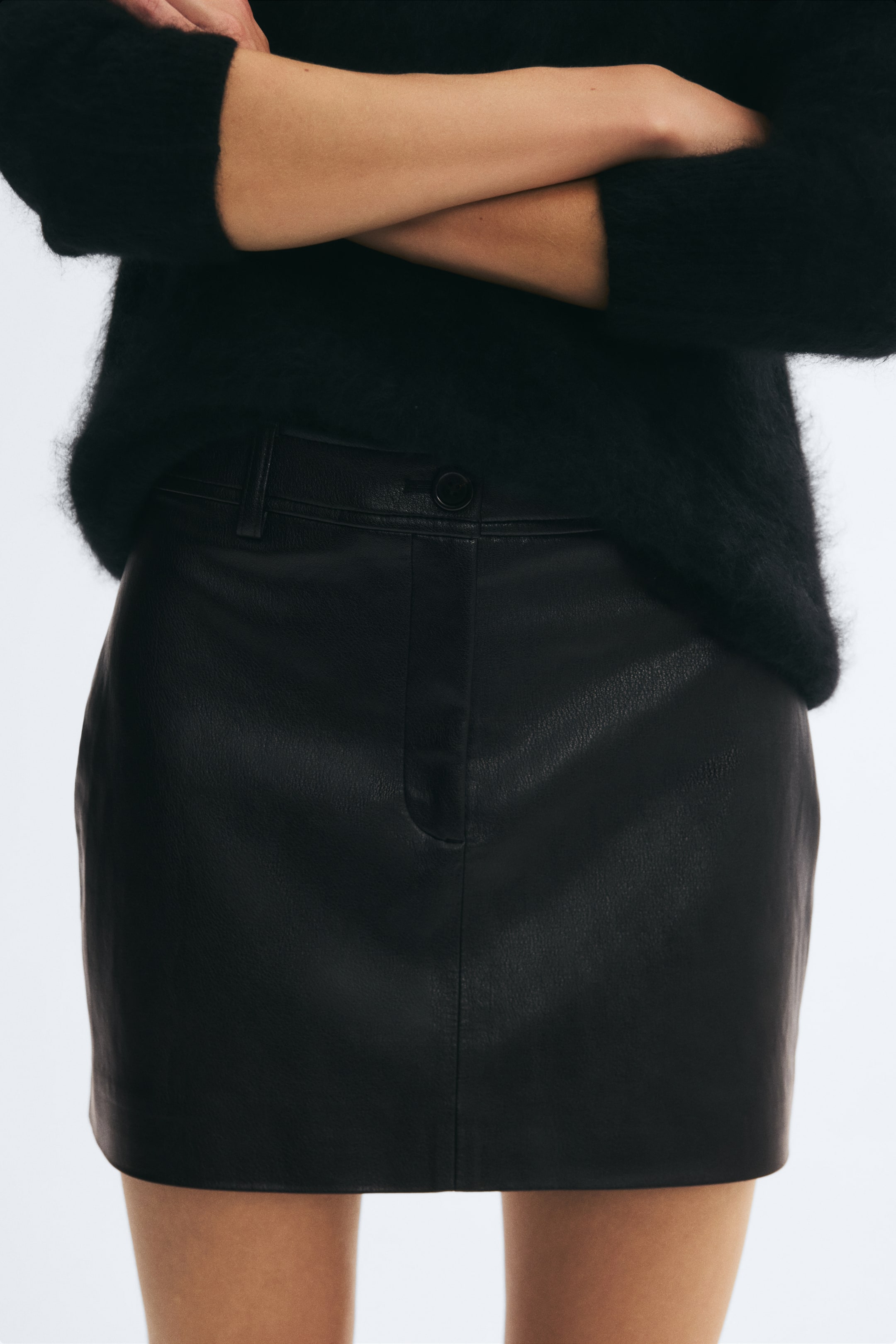 View larger image: The model is wearing a black leather mini skirt with a button closure, zip fly, and belt loops. It is styled with a fuzzy black long-sleeved sweater layered over the waistband.