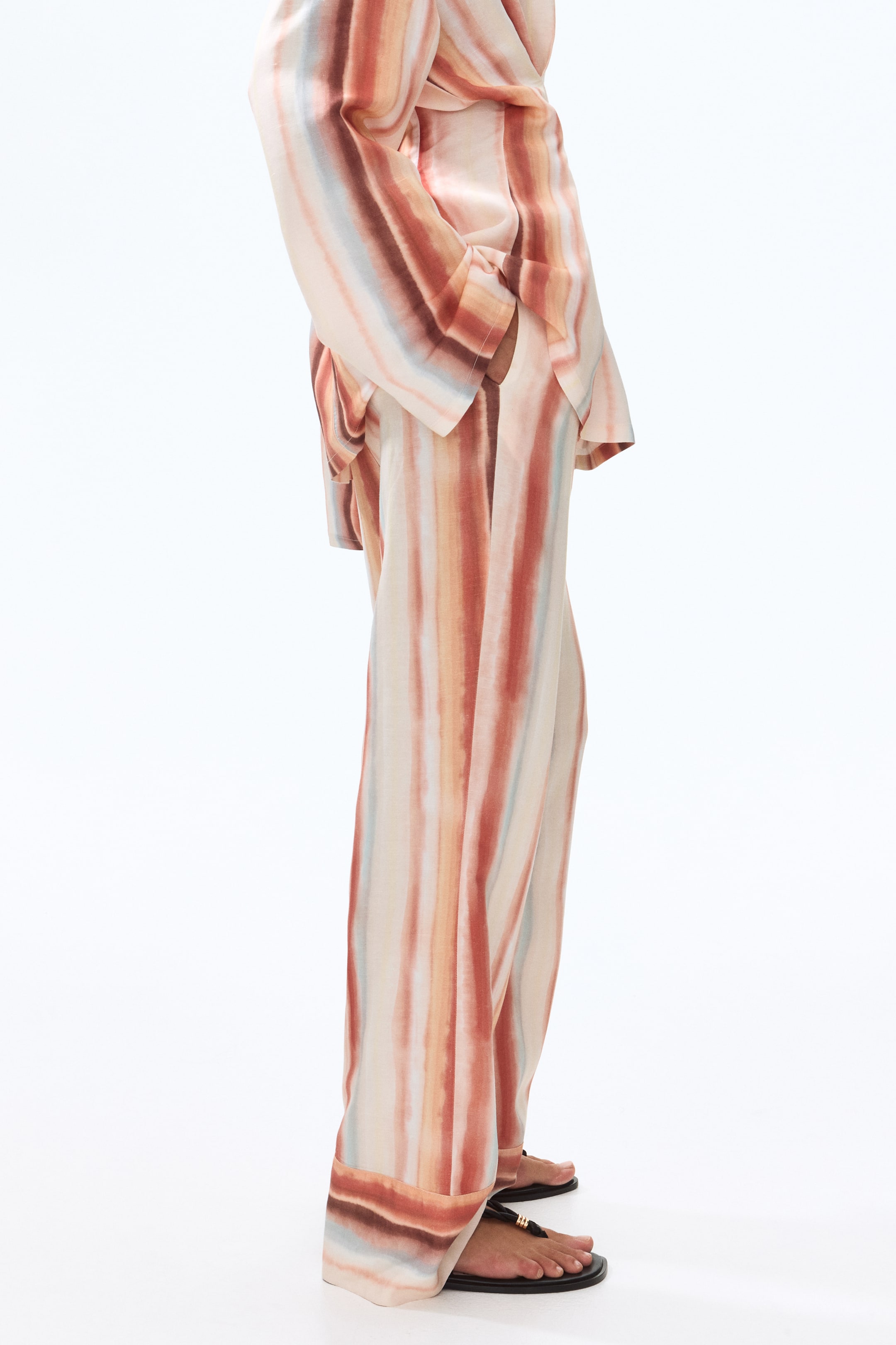 View larger image: Loose-fit trousers crafted from a breathable weave, combining linen and viscose, are showcased by the model. The trousers feature vertical watercolor stripes in hues of brown, orange, peach, and light blue. The model is seen with one hand casually placed in a subtle side pocket, while the wide-legged design is finished with double-layered borders at the hems.