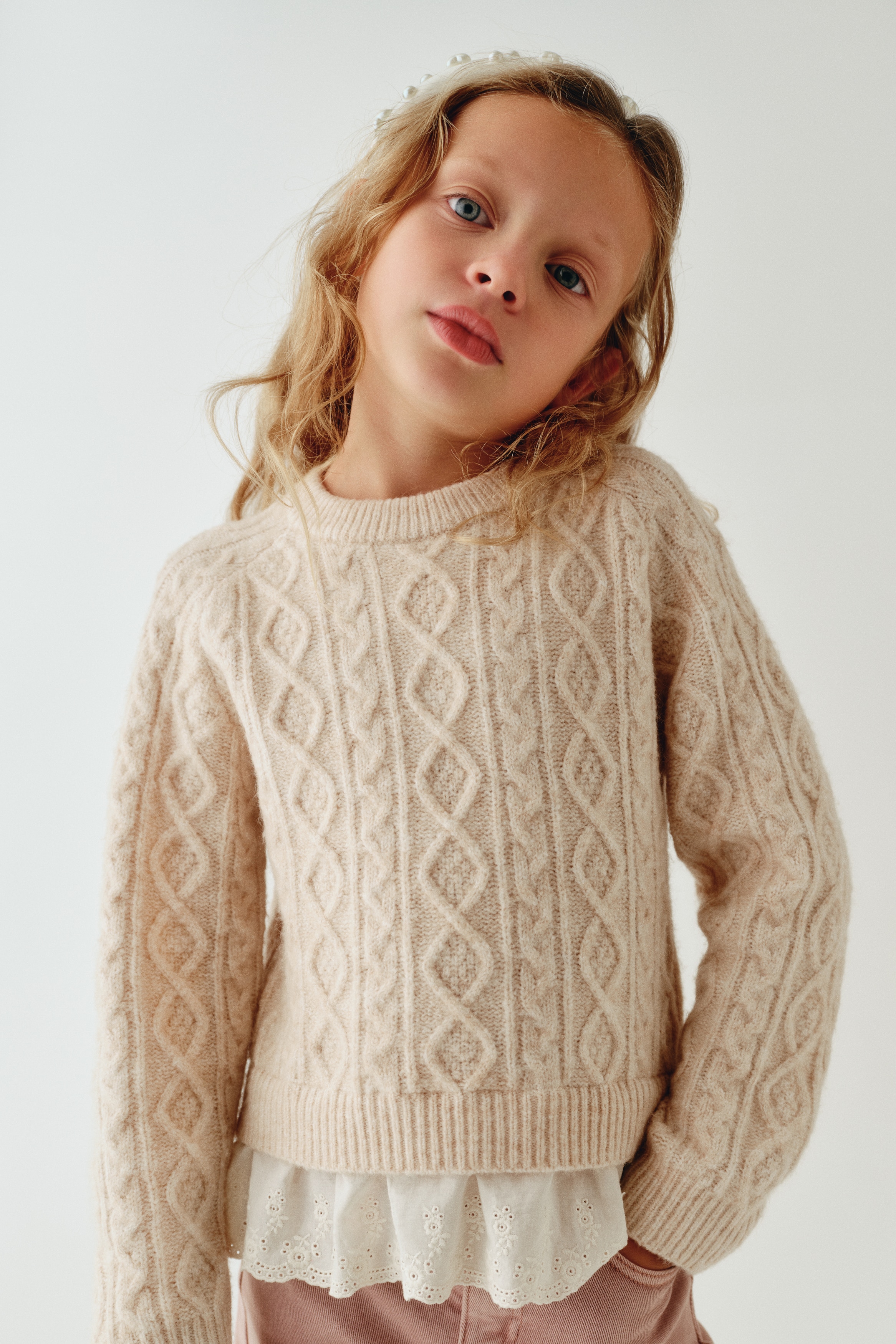 A cream-coloured cable-knit sweater is shown from the front, featuring a ribbed crew neck, ribbed cuffs, and a ribbed hem. A white ruffled eyelet-lace underlayer peeks out from beneath the bottom hem, creating a layered look.