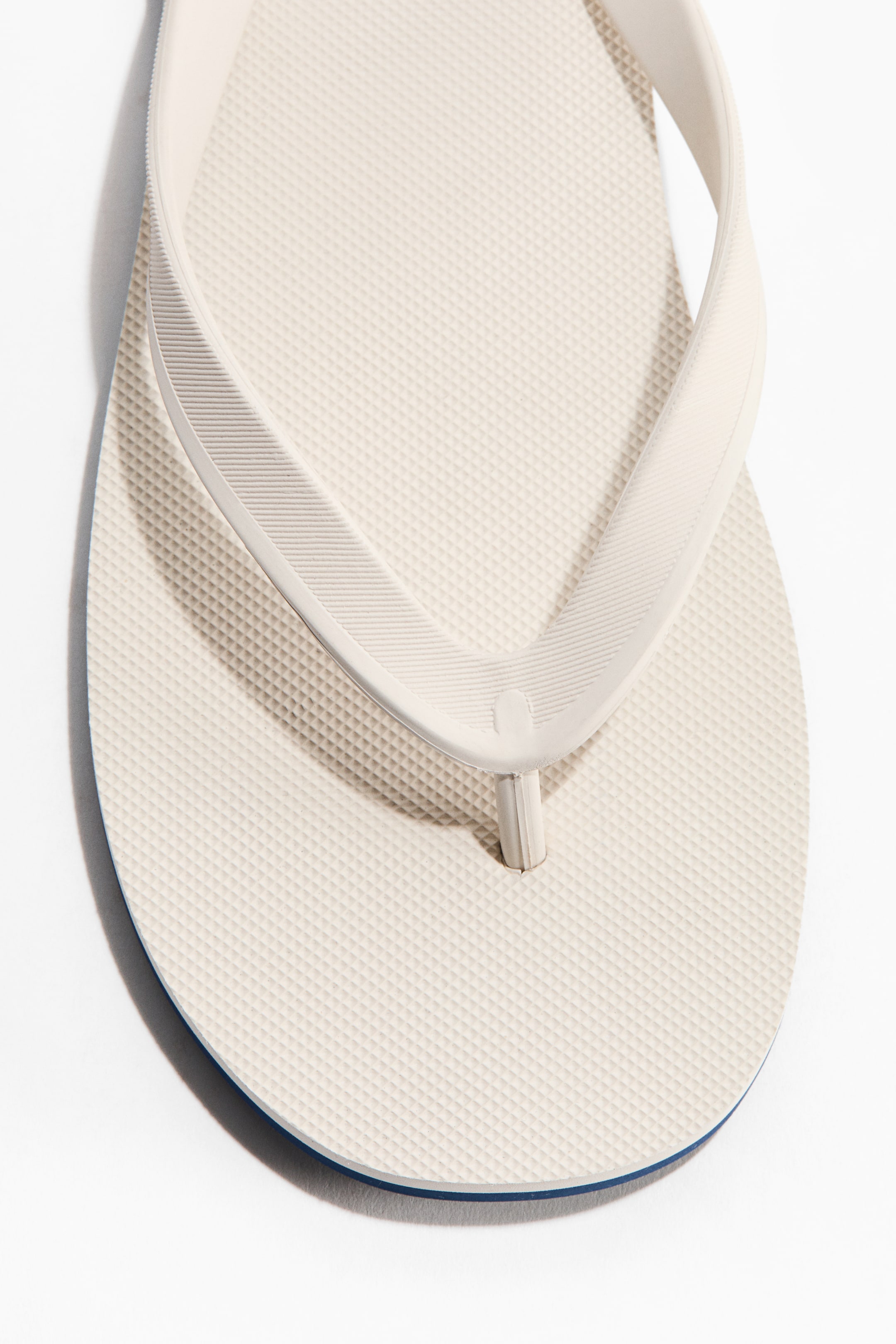 View larger image: A cream-colored rubber flip-flop is showcased from the front, highlighting straps with a ribbed inner surface that converge into a narrow toe post. The footbed displays a diamond-patterned texture, while a thin blue line outlines the sole.