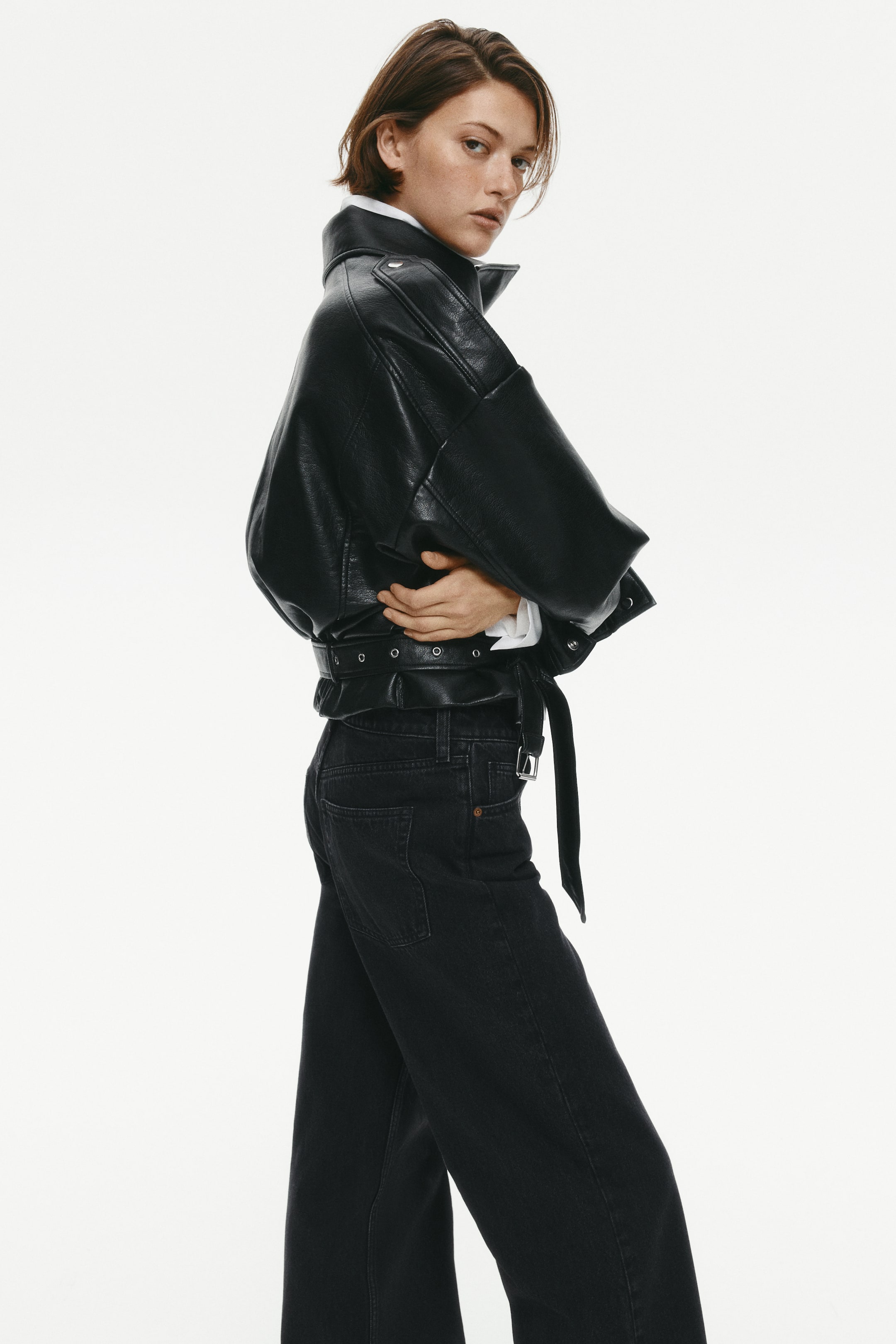 View larger image: The model is wearing black wide-leg denim jeans, featuring a regular waist, a classic back pocket, and a loose, baggy fit through the legs. The jeans, styled with a matching black belt, are paired with a black leather jacket and a white shirt.