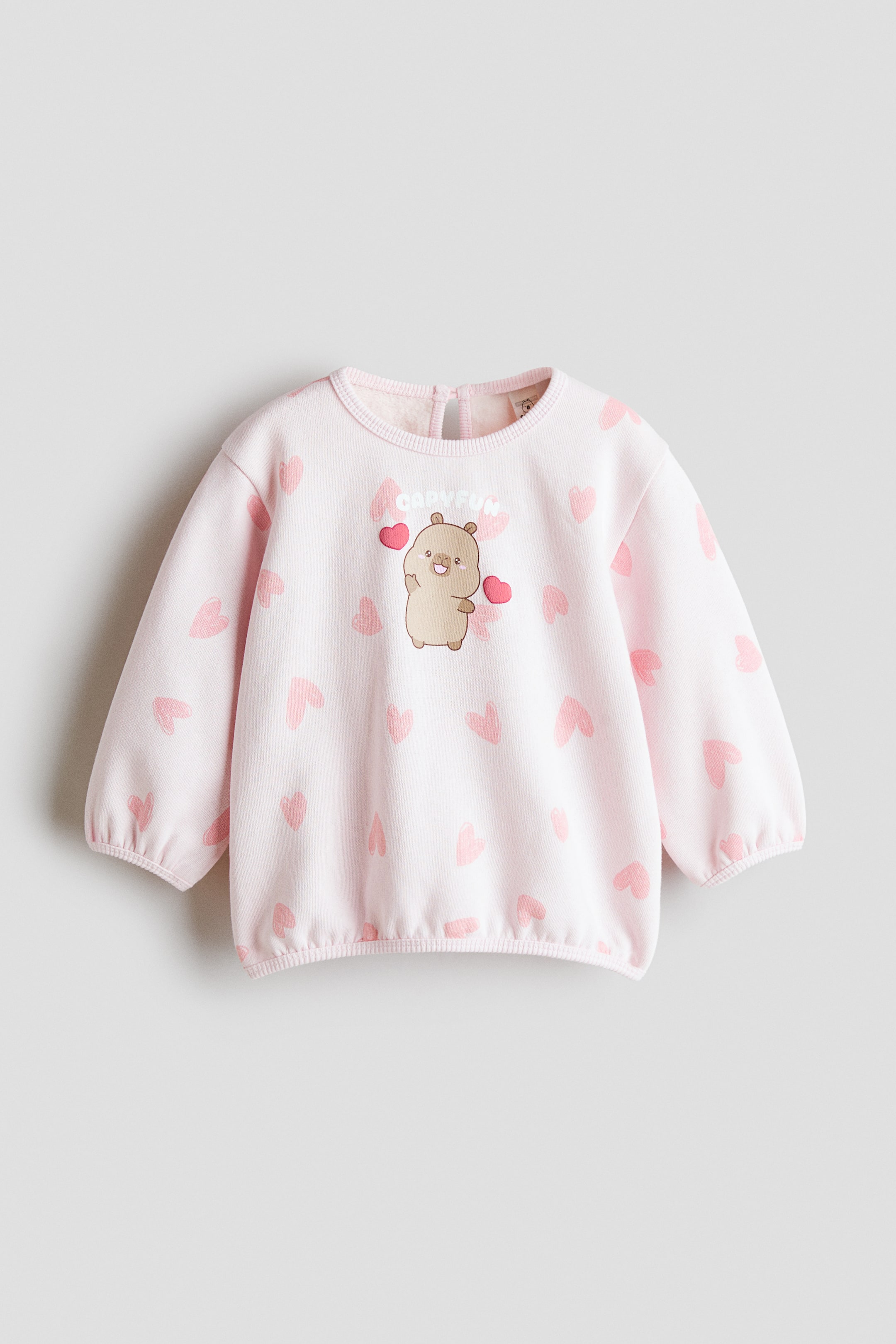 Ver imagem maior: Uma sweatshirt rosa-claro de manga comprida está voltada para a frente, apresentando um padrão integral de pequenos corações rosa e um gráfico central em estilo cartoon de um capivara castanha com a palavra "CAPYFUN" impressa por cima. A sweatshirt exibe gola redonda canelada, punhos elásticos e bainha com elástico franzido.