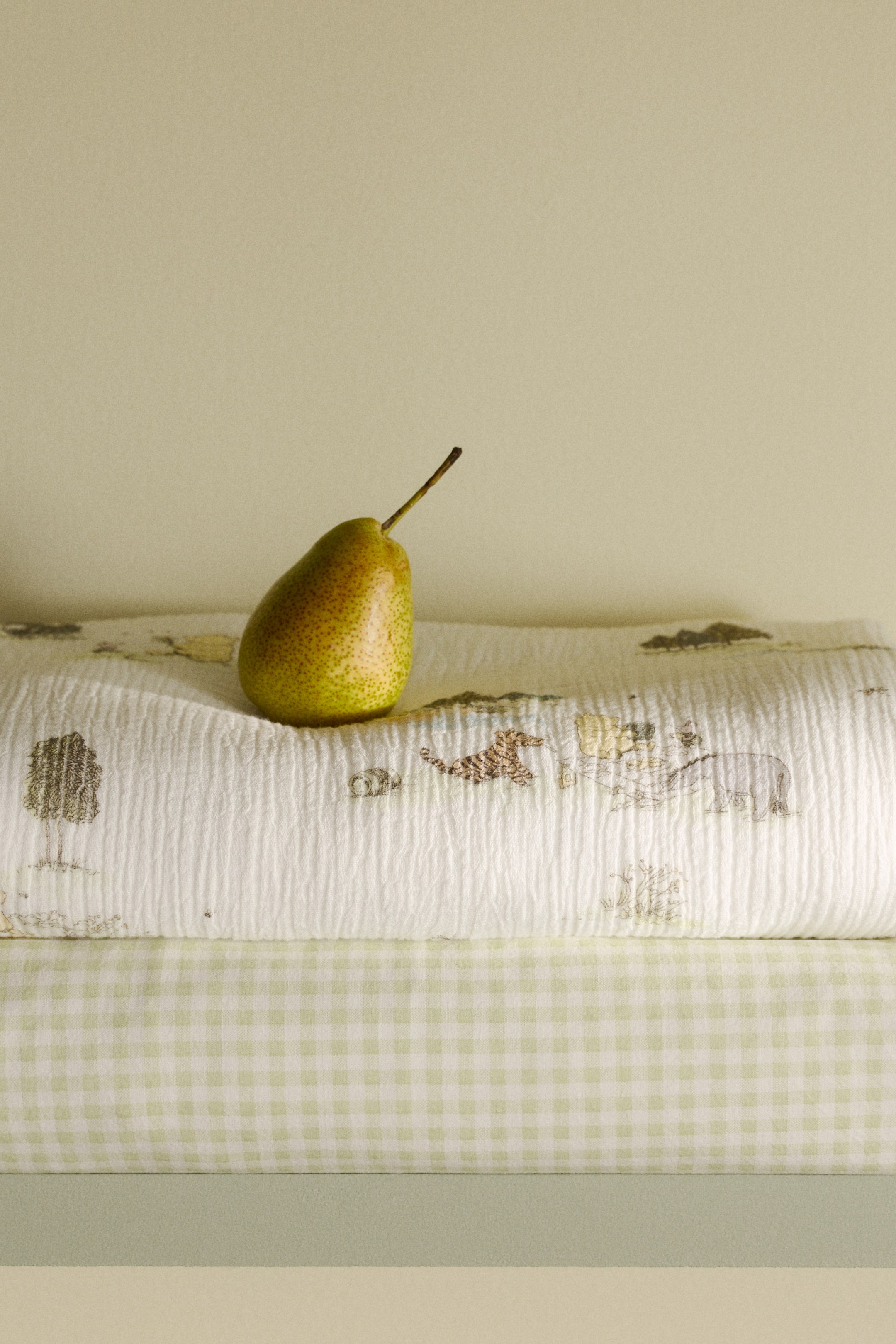 View larger image: A greenish-yellow pear is standing upright, showcasing its speckled skin and a brown stem protruding from its top. It is set on a white, crinkled fabric adorned with subtle patterns of trees and animals, layered over a light green gingham fabric.