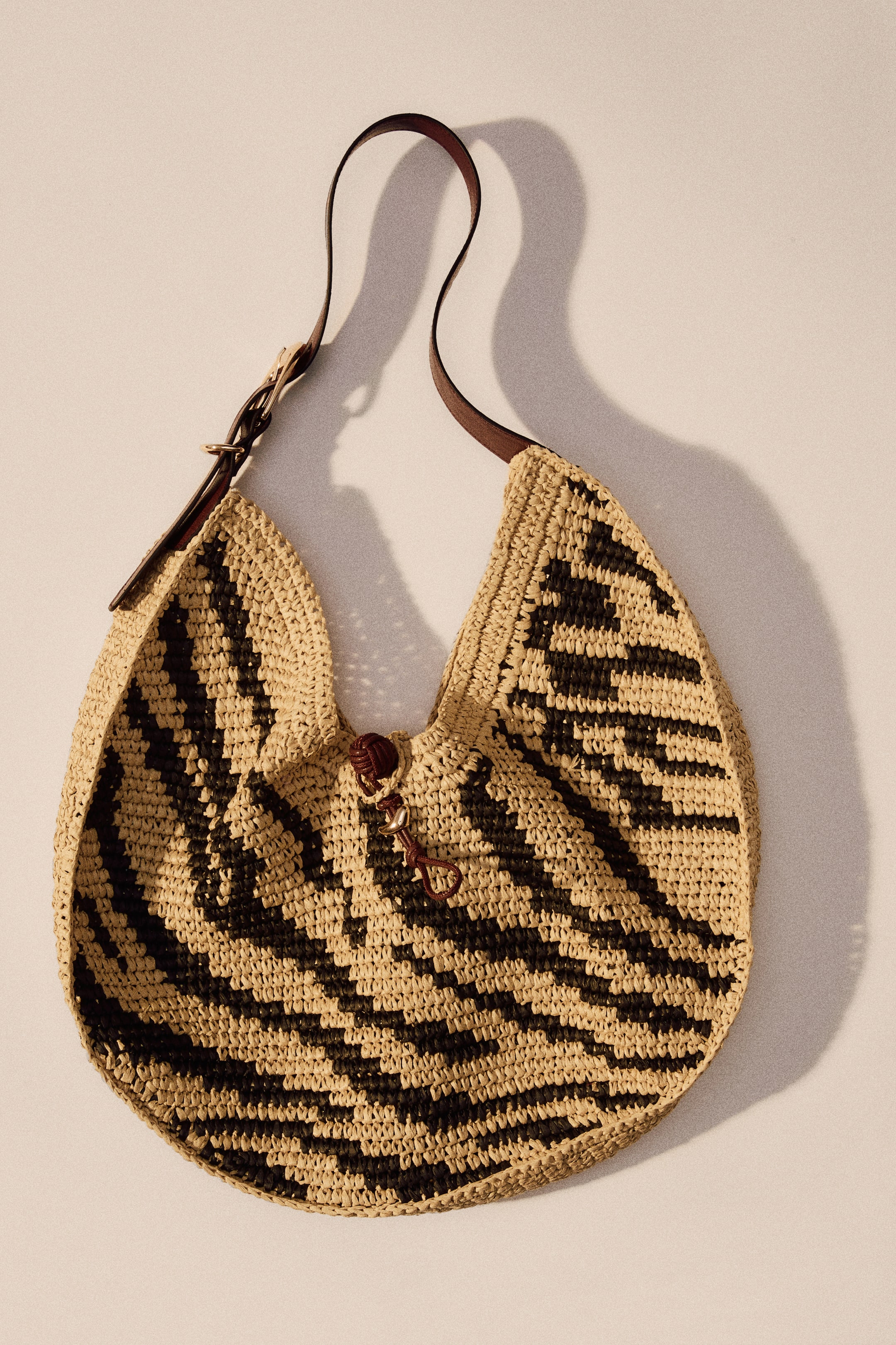 Agrandir l'image: Un sac à bandoulière en paille de papier tressée présente un motif géométrique noir et beige, présenté de face. Il est doté d'une bandoulière réglable en tissu enduit marron foncé avec une boucle dorée sur un côté, et un fermoir à bouton et boucle noué marron foncé avec un petit charme en métal doré sécurise l'ouverture supérieure.