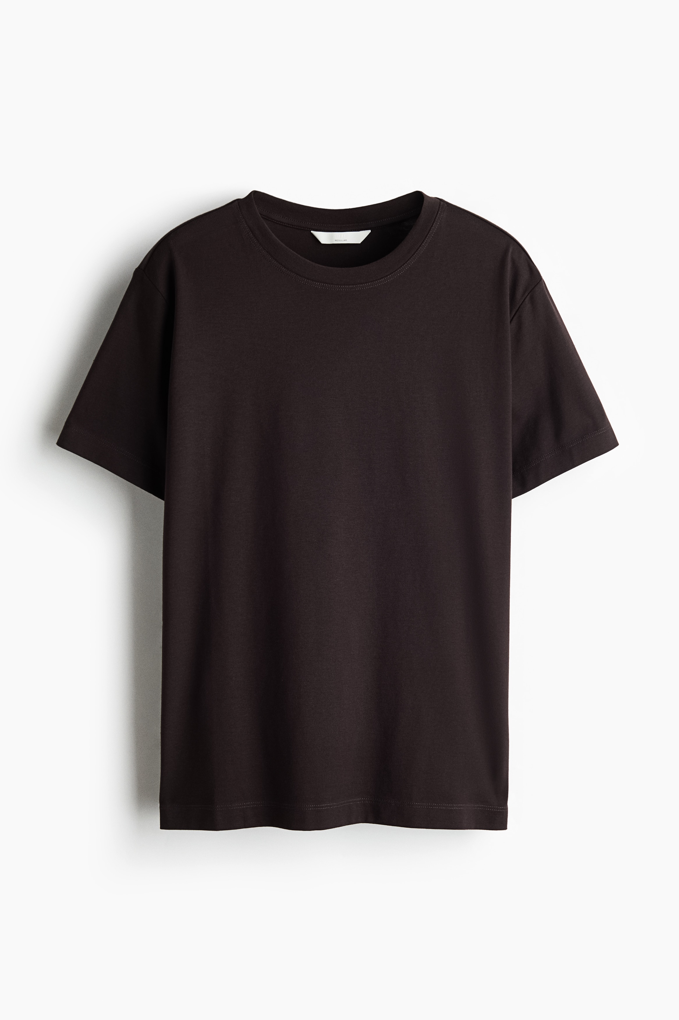 Een donkerbruine T-shirt met ronde hals en korte mouwen wordt gedragen door het model en gecombineerd met een witte jeans met hoge taille. Het model staat met één hand in een broekzak, waardoor de relaxte pasvorm van het topje goed zichtbaar is.