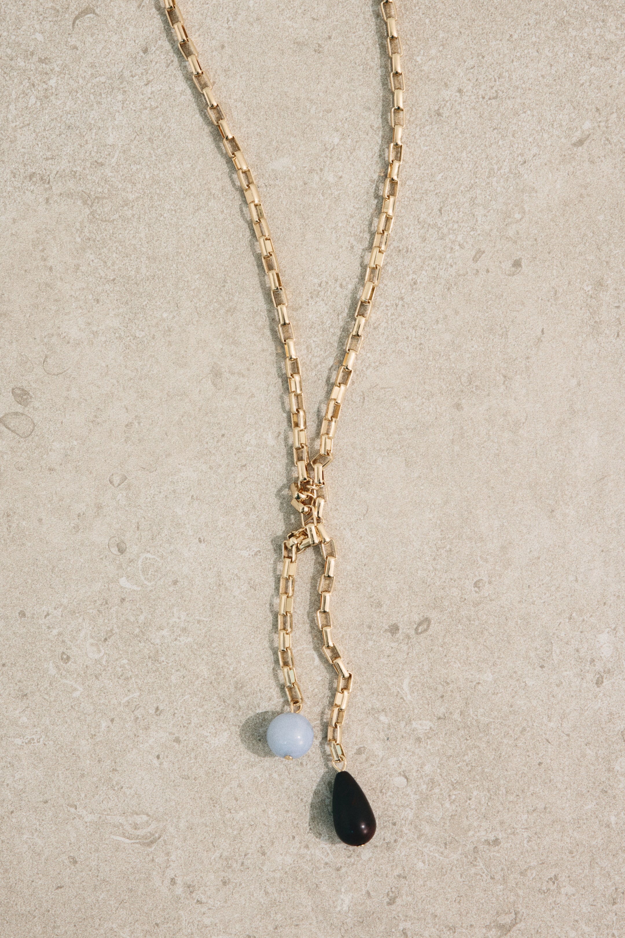 Grösseres Bild ansehen: Eine goldfarbene Box-Ketten-Lariat-Halskette liegt flach und zeigt einen zentralen Knoten, von dem eine hellblaue kugelförmige Perle und eine dunkle tropfenförmige Perle hängen.