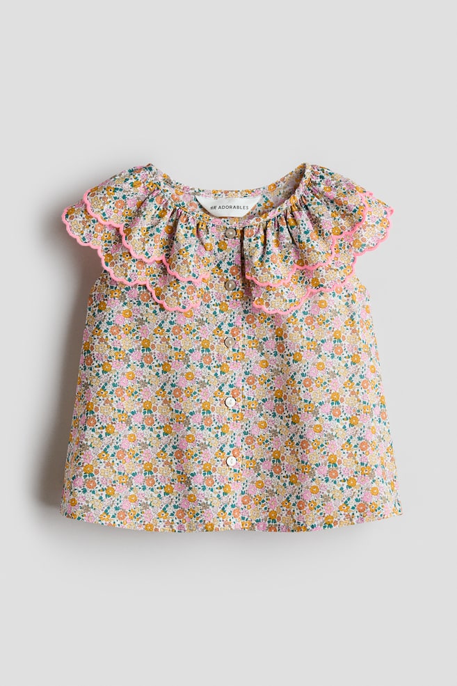 Se muestra una blusa sin mangas de H&M Adorables desde el frente, con un pequeño estampado floral en rosa, amarillo y verde, un escote redondo y una tapeta de botones en la parte delantera. Su cuello con doble capa de volantes presenta bordes festoneados con sobrehilado rosa. - 1