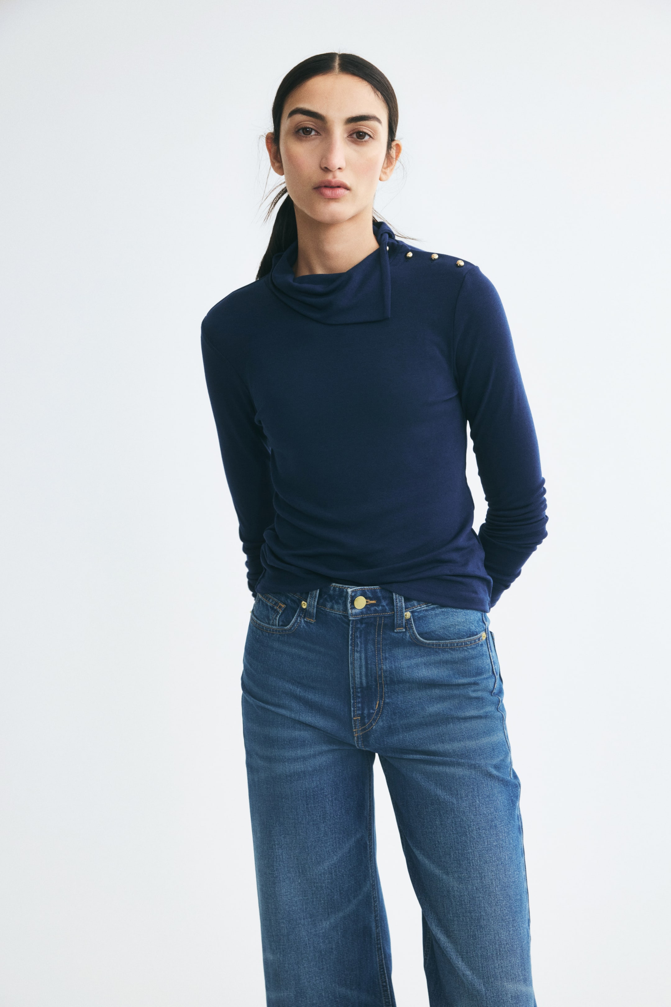 View larger image: A dark blue, fitted jersey top featuring an asymmetrical draped turtleneck and gold spherical buttons on one shoulder is being worn by the model. The long-sleeved top is paired with high-waisted blue jeans.