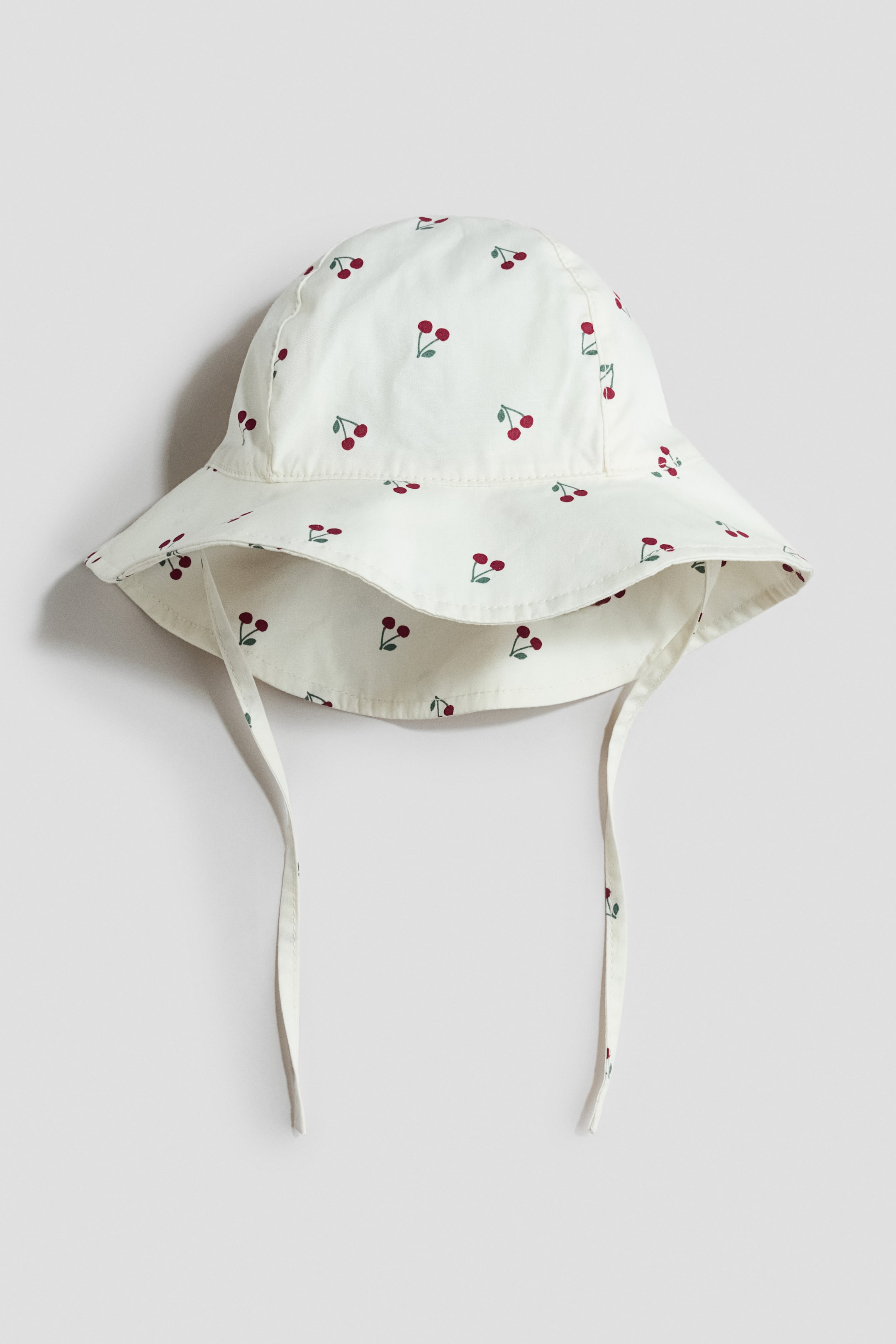 View larger image: A cream sun hat showcases a delightful pattern of petite red cherries. Its brim elegantly slopes downwards, with ties extending gracefully from beneath the chin.
