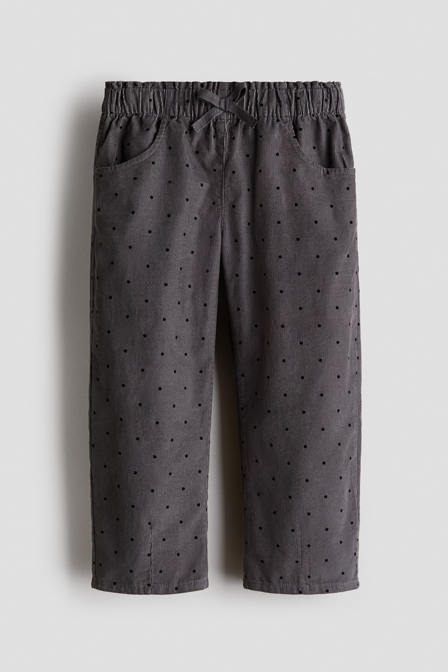 A pair of dark grey corduroy pants is shown from the front, featuring an all-over pattern of small black polka dots. The pants have a gathered elastic waistband with a decorative matching bow at the centre and two curved side pockets. - 1