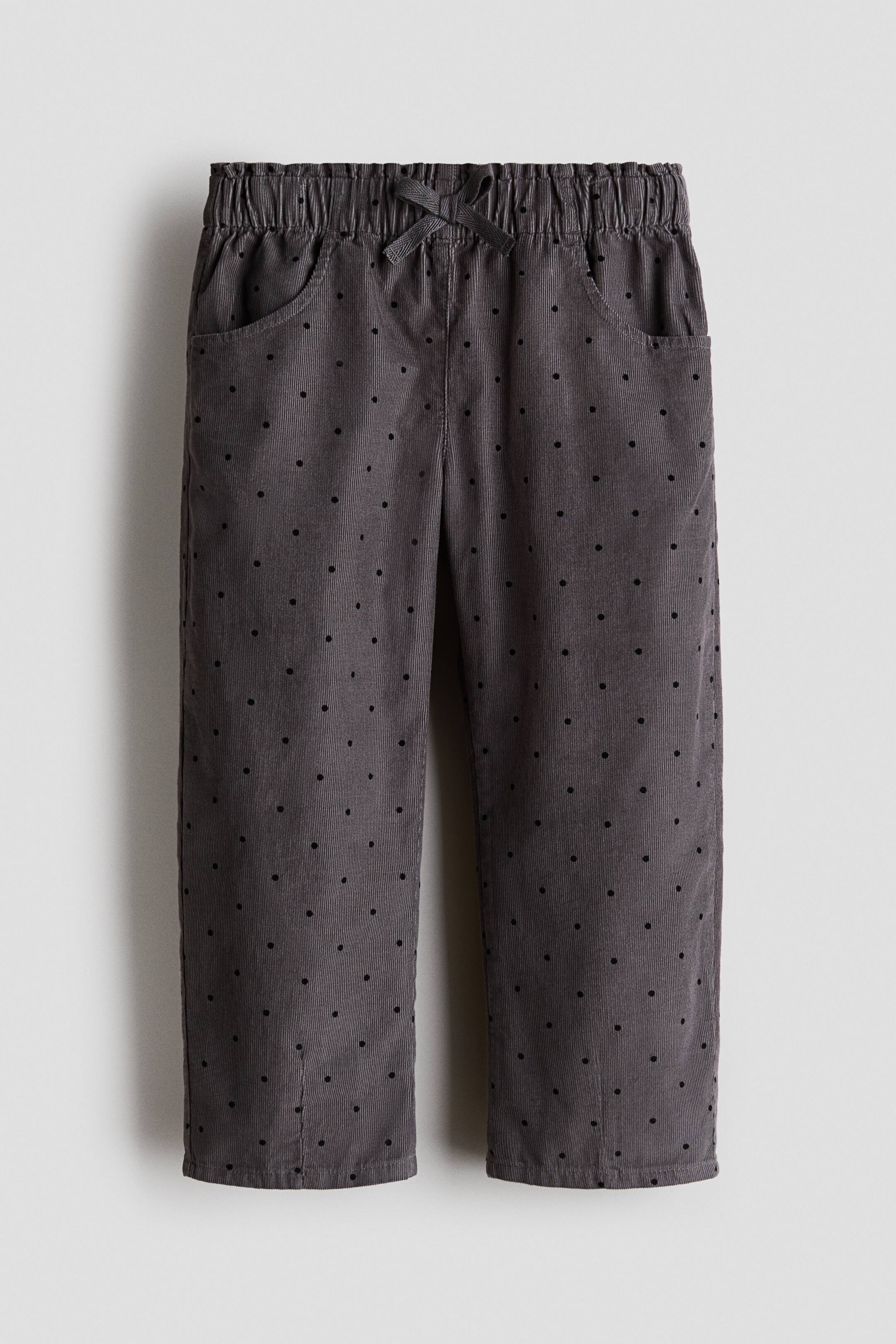 View larger image: A pair of dark grey corduroy pants is facing front, featuring an all-over pattern of small black polka dots. The pants have a gathered elastic waistband with a decorative matching bow at the centre and two curved side pockets.