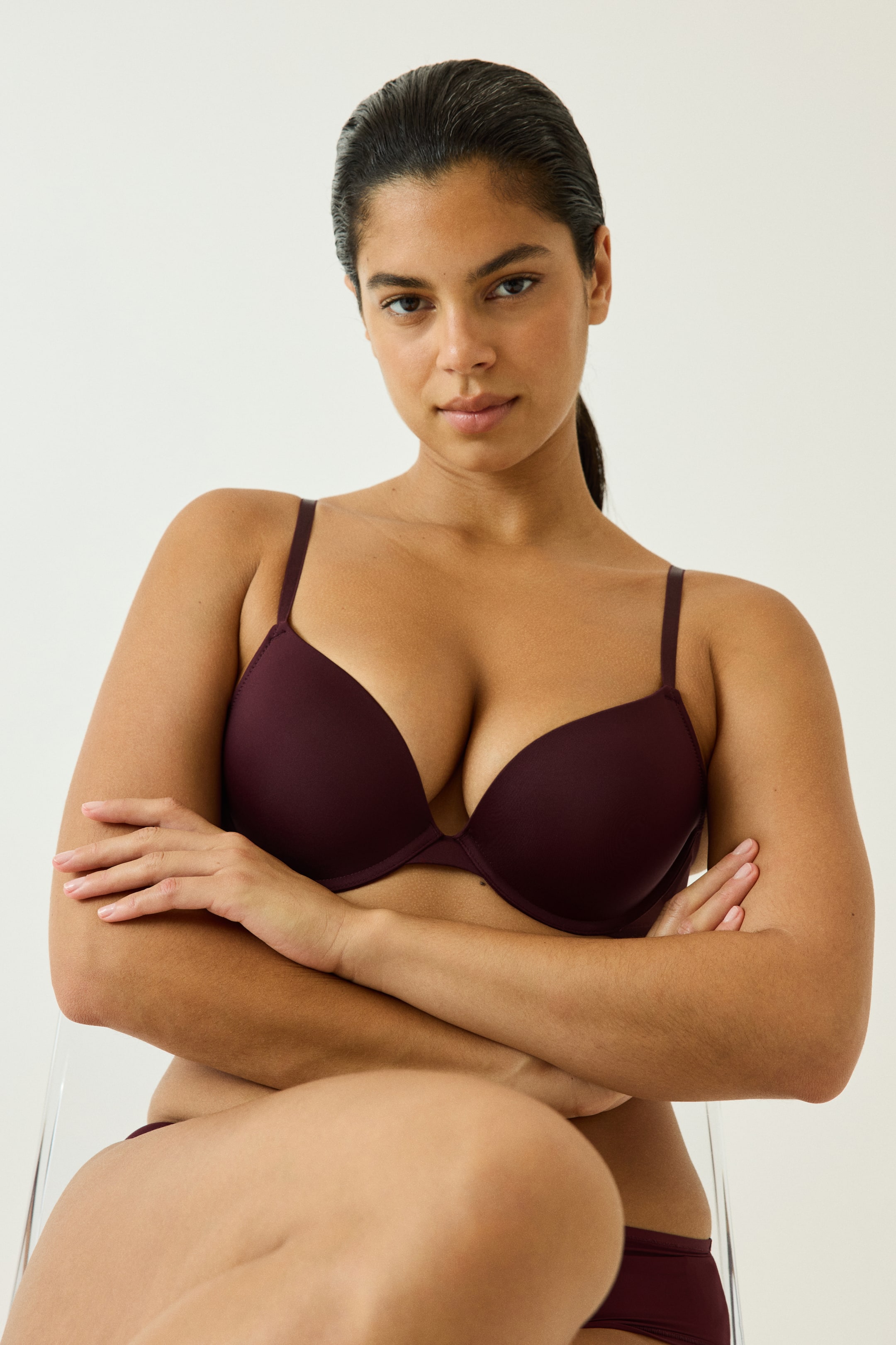 View larger image: A dark plum microfibre push-up bra featuring padded, underwired cups and slim shoulder straps is being worn by the model. The model is sitting with folded arms, wearing the bra alongside matching briefs.