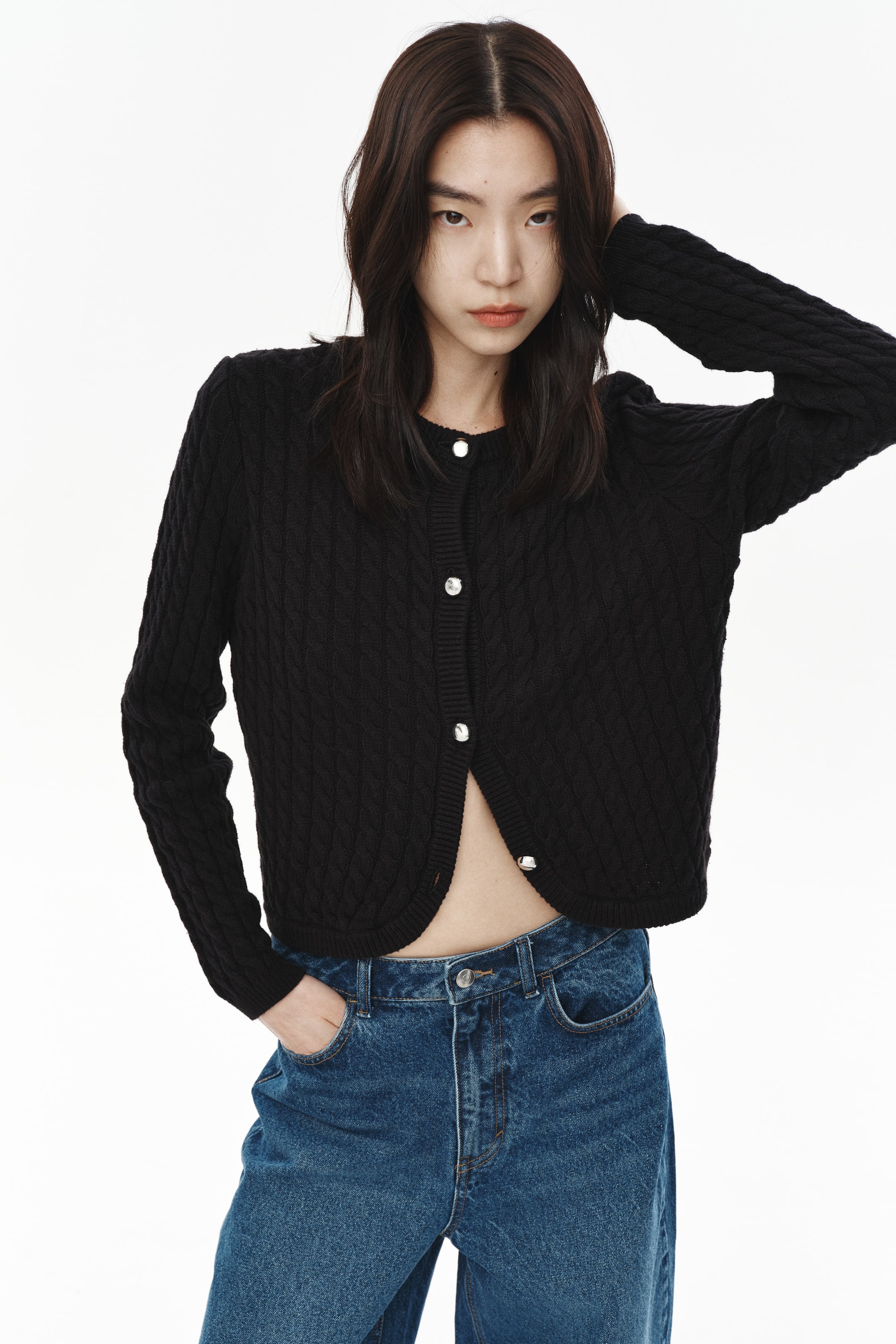 이미지 확대 보기: 앞면에서 보여지는 복잡한 니트 패턴이 있는 검은색 가디건입니다. 둥근 목선, 긴 소매, 그리고 앞면의 다섯 개의 은색 버튼이 보이는 버튼 클로저가 특징입니다. 상단 두 개의 버튼은 잠겨 있고, 하단 세 개는 풀려 있어 크롭 핏과 곡선형 밑단을 강조합니다.