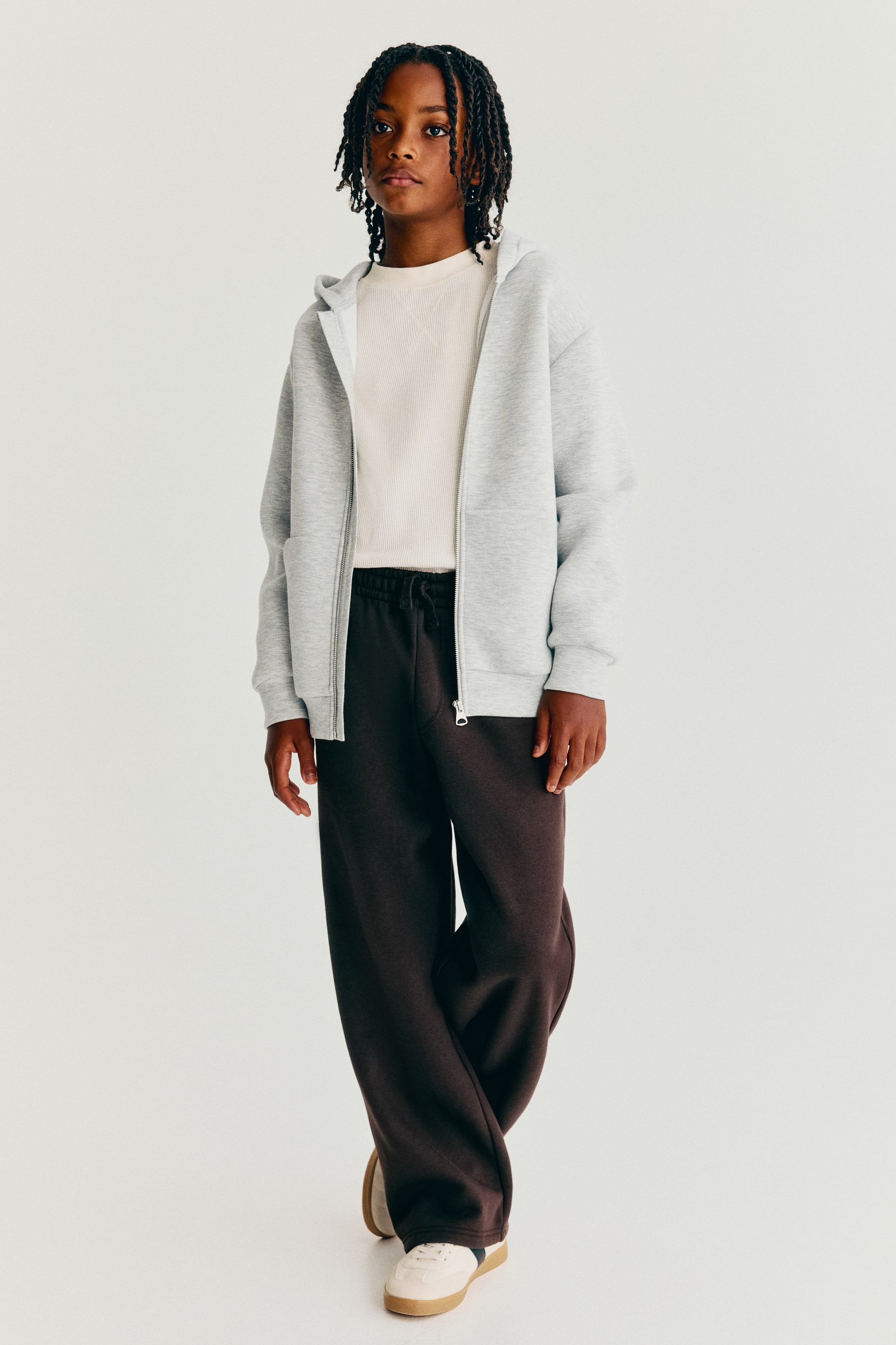 Agrandir l'image: Un pantalon de survêtement marron foncé est présenté de face, avec une silhouette ample à jambe large et une taille élastiquée avec cordon de serrage. Le pantalon tombe de façon fluide sur des baskets, associé à un hoodie zippé gris clair ouvert et à un t-shirt blanc.
