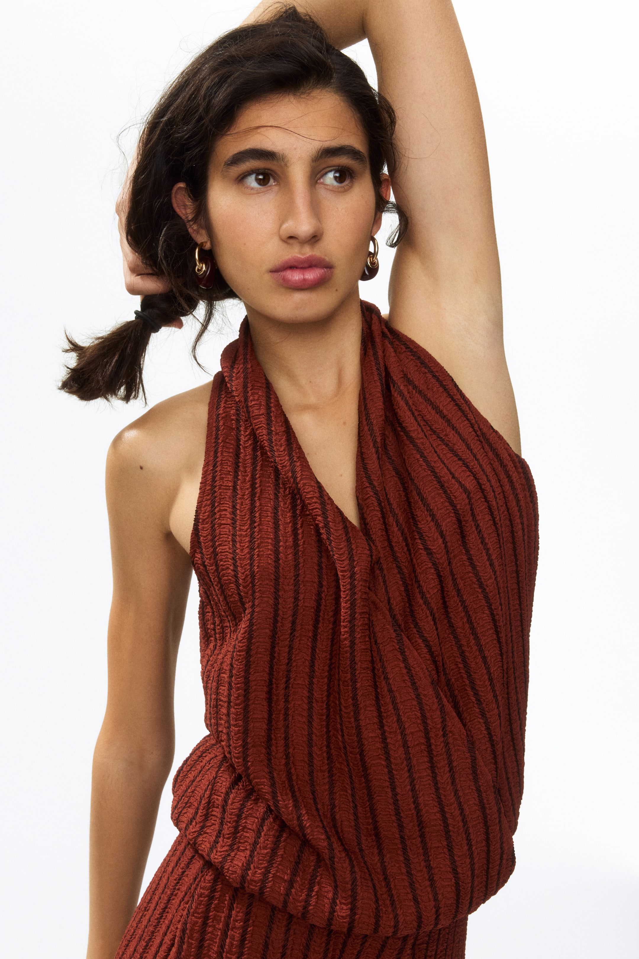 Agrandir l'image: Un haut à col halter de couleur rouille présente une texture froissée, formant de subtiles crêtes verticales, avec de fines rayures horizontales marron foncé. Il est conçu avec un décolleté en V profond et tombe librement sur le torse, se rassemblant autour de la taille.