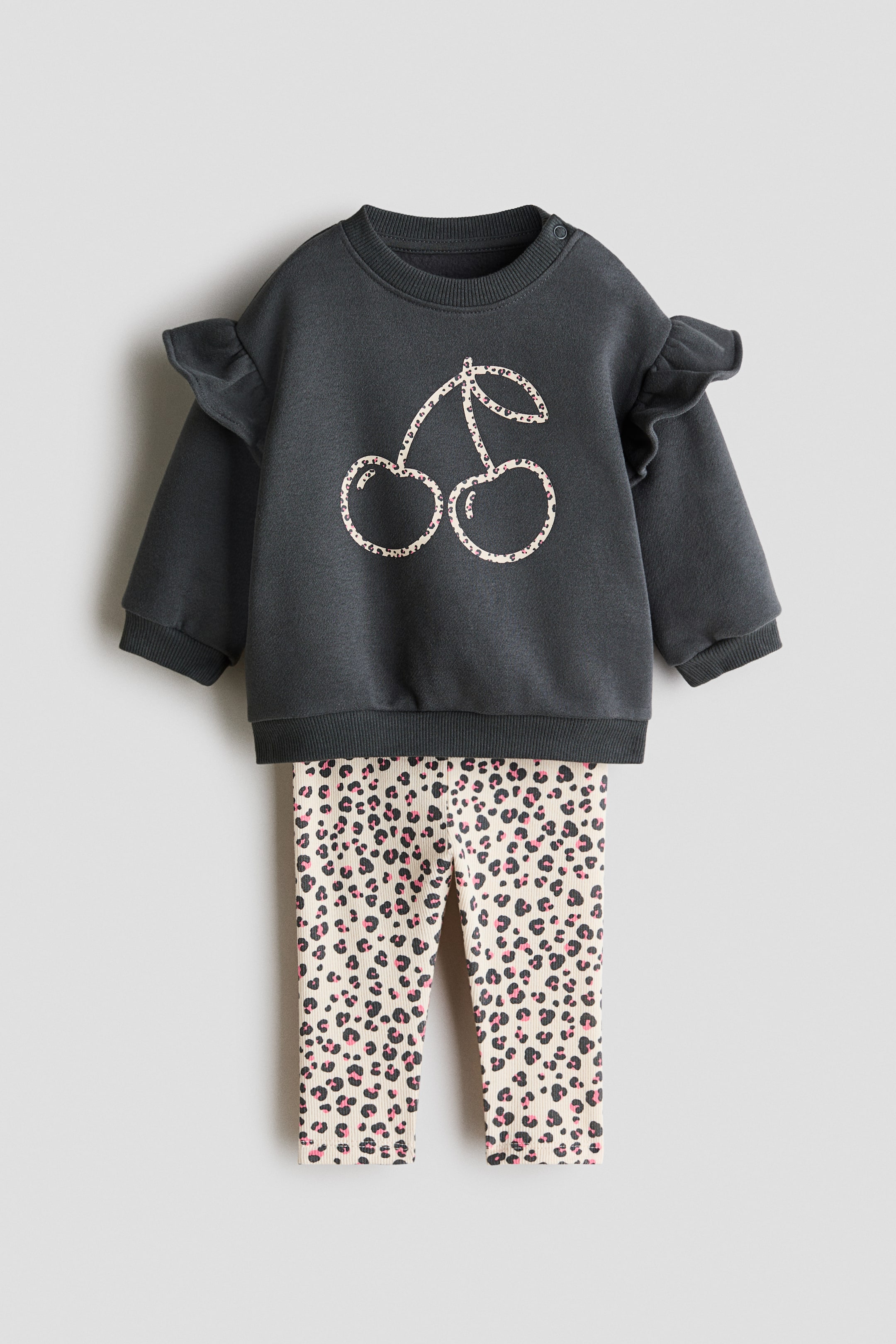 View larger image: An infant’s dark grey long-sleeved sweatshirt with ruffled shoulders, ribbed cuffs and hem, and a white-outlined leopard print cherry graphic on the front is paired with cream ribbed leggings featuring a black and pink leopard print. The two-piece outfit is shown front-facing.