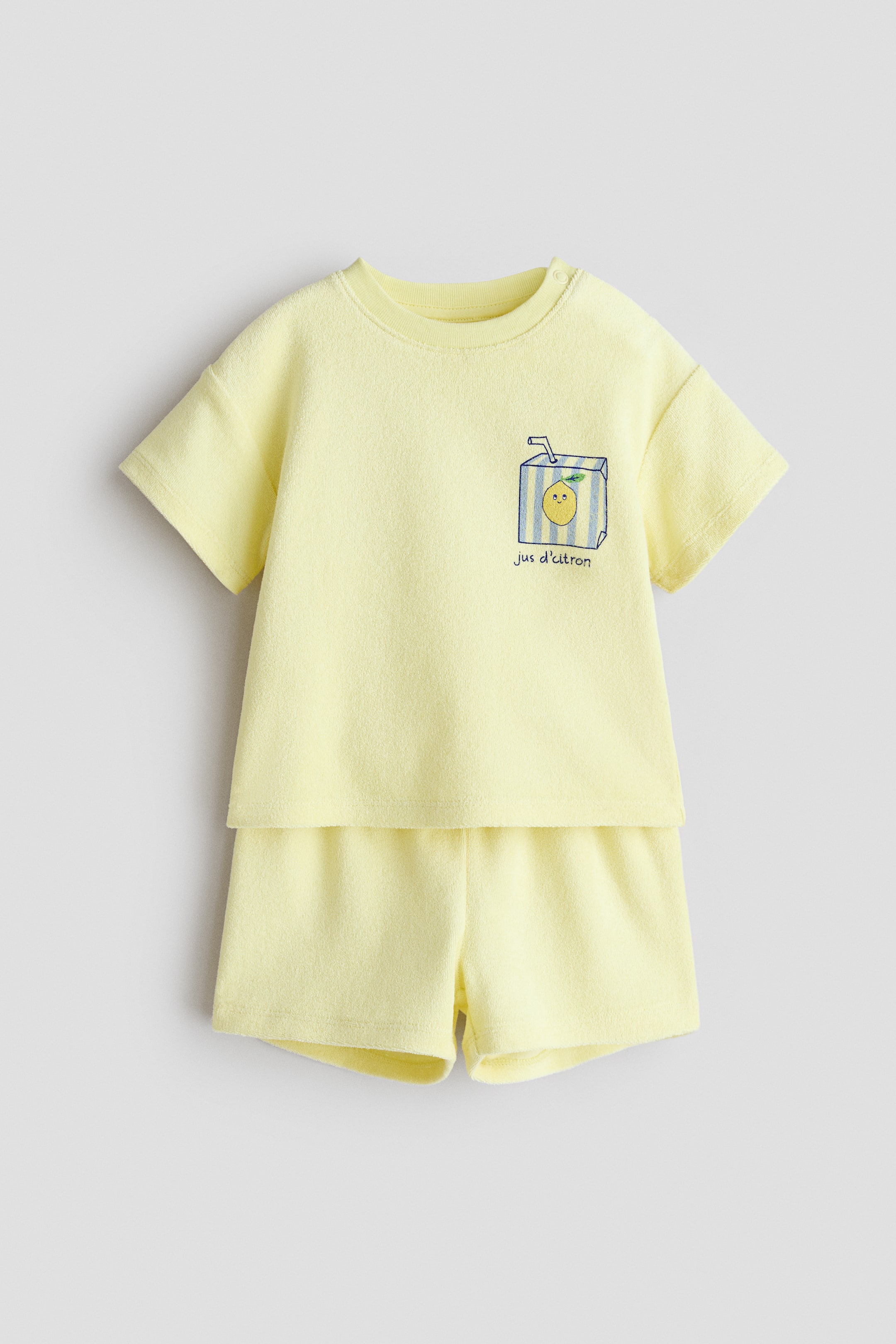 Agrandir l'image: Un ensemble bébé deux pièces jaune est présenté de face, comprenant un haut en tissu éponge à manches courtes et un short assorti. Le haut présente un col rond avec des boutons-pression à l'épaule gauche et un graphique d'une boîte de jus rayée avec un visage de citron et le texte "jus d'citron" sur la poitrine.
